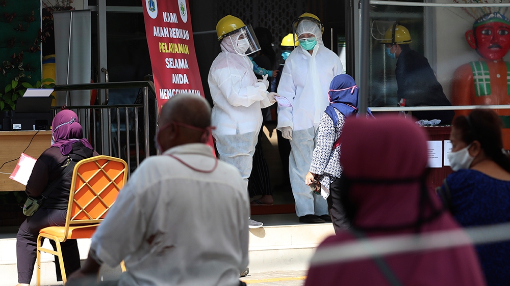 Staff in protective suits talk to a woman as people sit spaced apart to practice social distancing while waiting for their turn to enter a community health centre in Jakarta, Indonesia