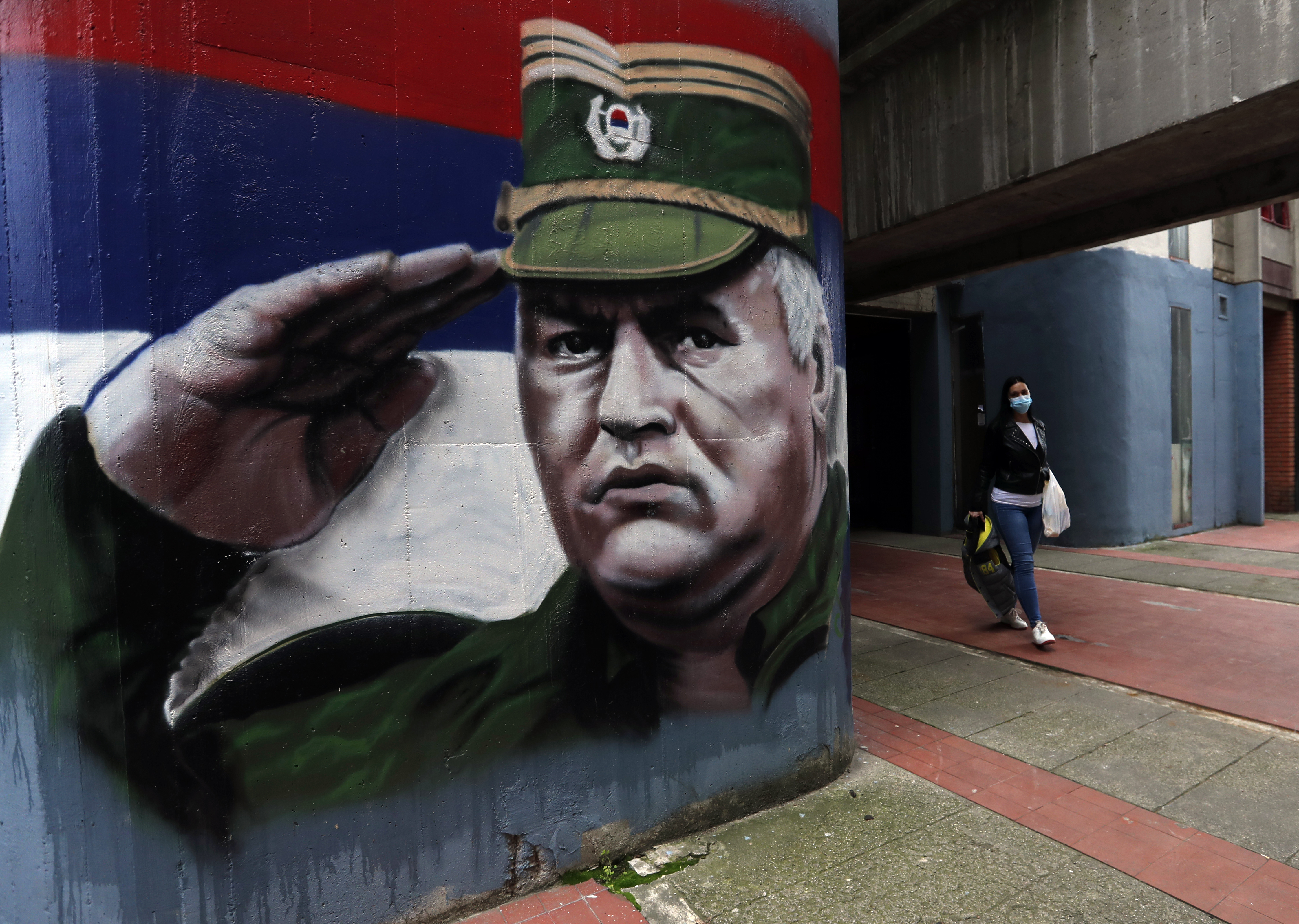 A woman wearing a face mask walks past graffiti depicting former Bosnian Serb wartime general Ratko Mladic in Belgrade, Serbia, Thursday, Nov. 12, 2020. Mladic was convicted by a UN war crimes tribunal in 2017 and sentenced to life imprisonment for masterminding crimes by Bosnian Serb forces throughout the war that left 100,000 dead. (AP Photo/Darko Vojinovic)