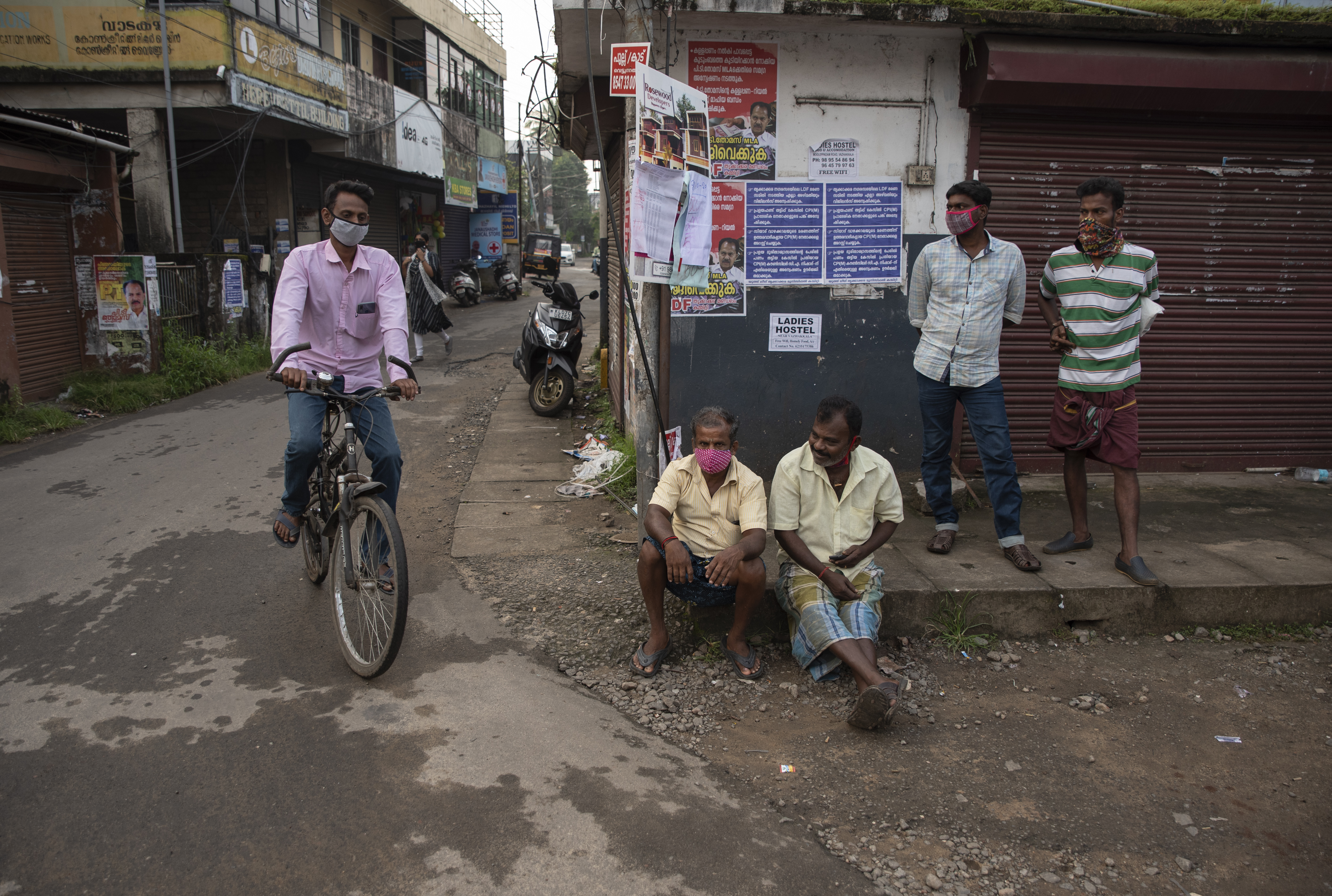 A cyclist wearing a face mask pedals past daily wage labourers waiting to be hired for the day, by a roadside in Kochi