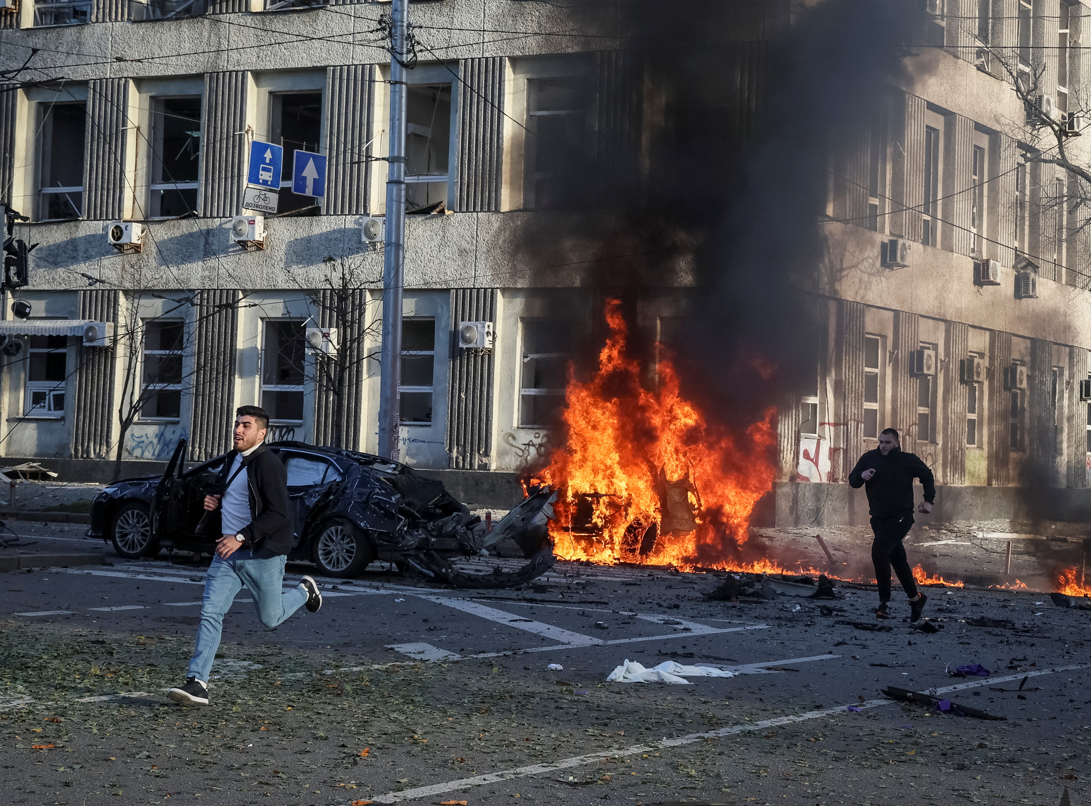 Two people run across the road, past the wreck of a car still burning, with plumes of black smoke rising over it