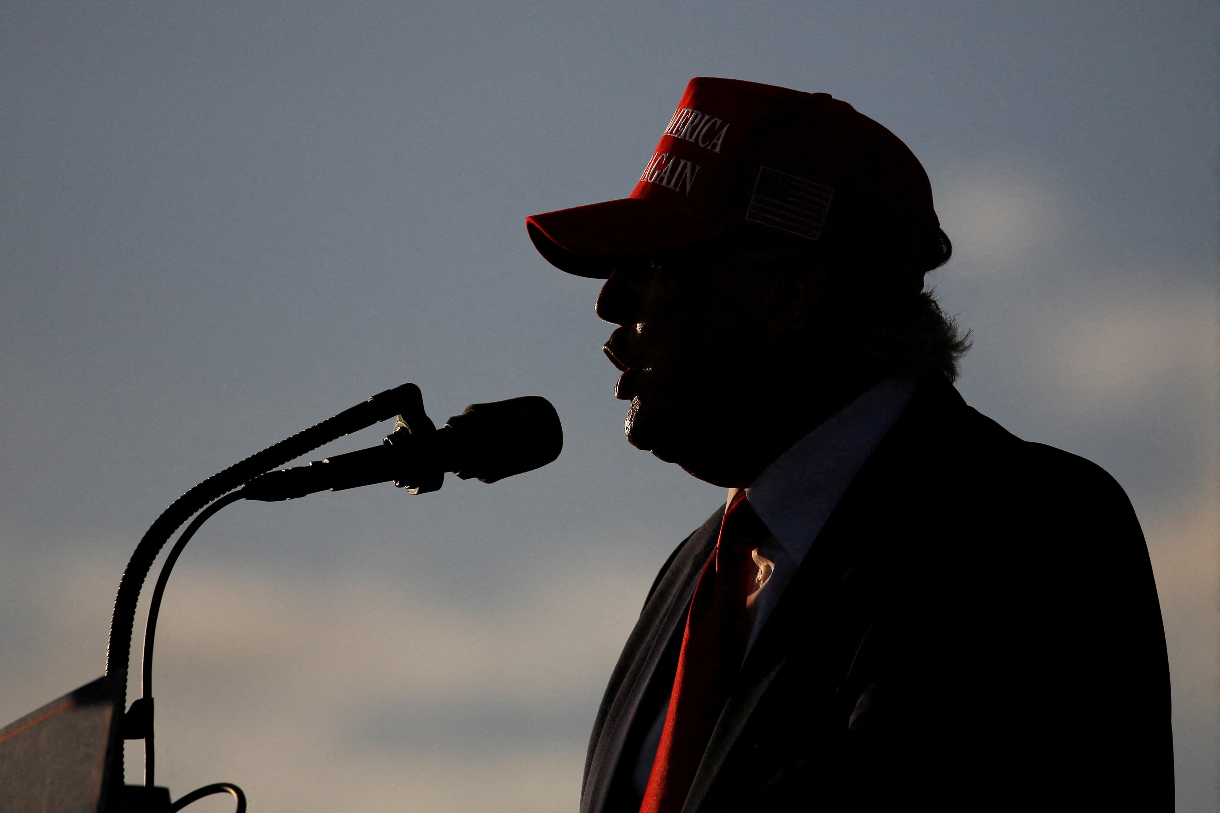 Donald Trump at an outdoor lectern with a hat on, silhouetted against the a blue sky with wisps of clouds.
