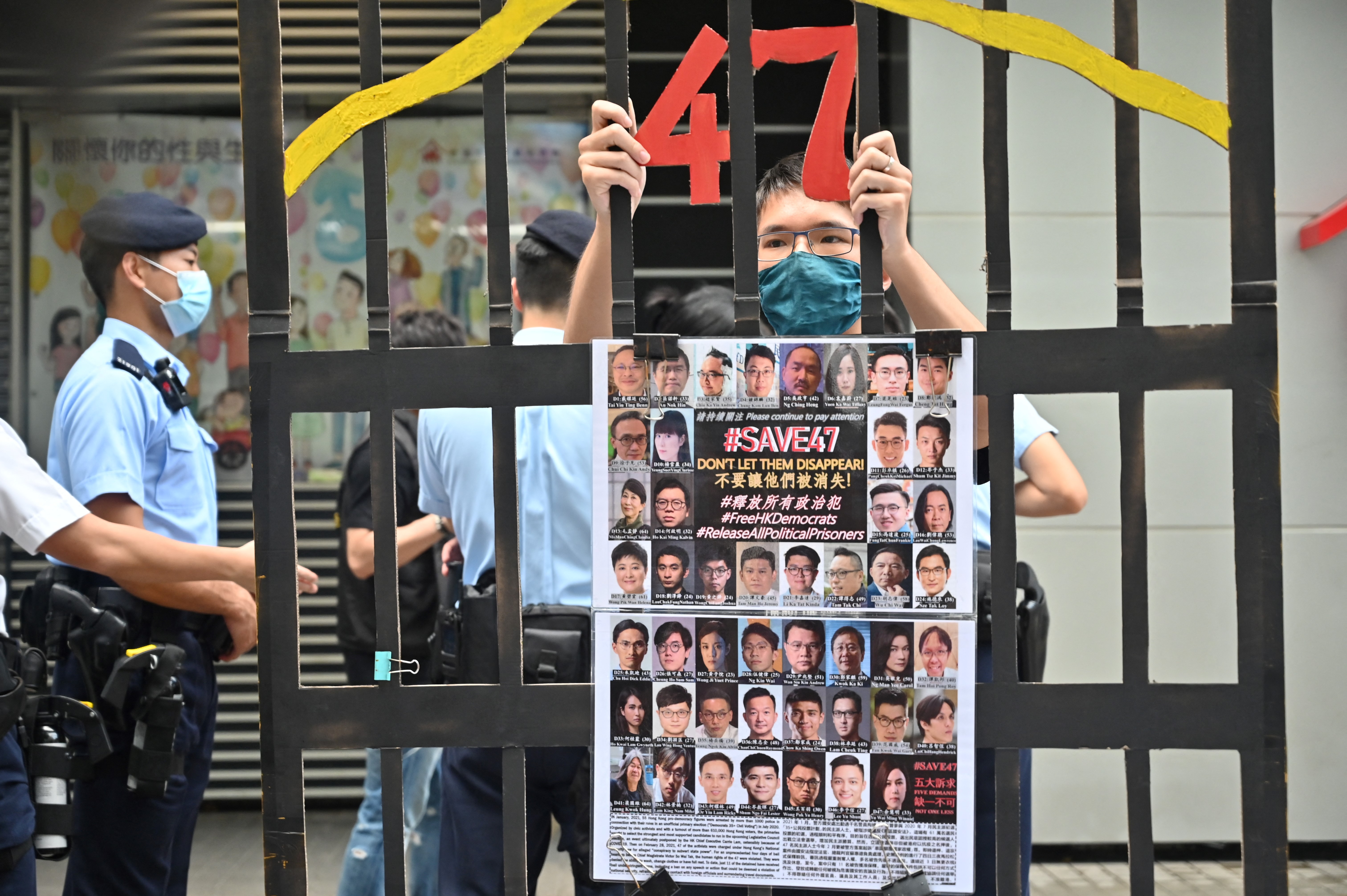 A man stands behind a mock-up of the bars of a jail cell holding a banner showing the 47 Hong Kong activists and legislators charged under security laws for arranging their own primary. The number 47 is in red at the top of the bars. There are masked police officers behind