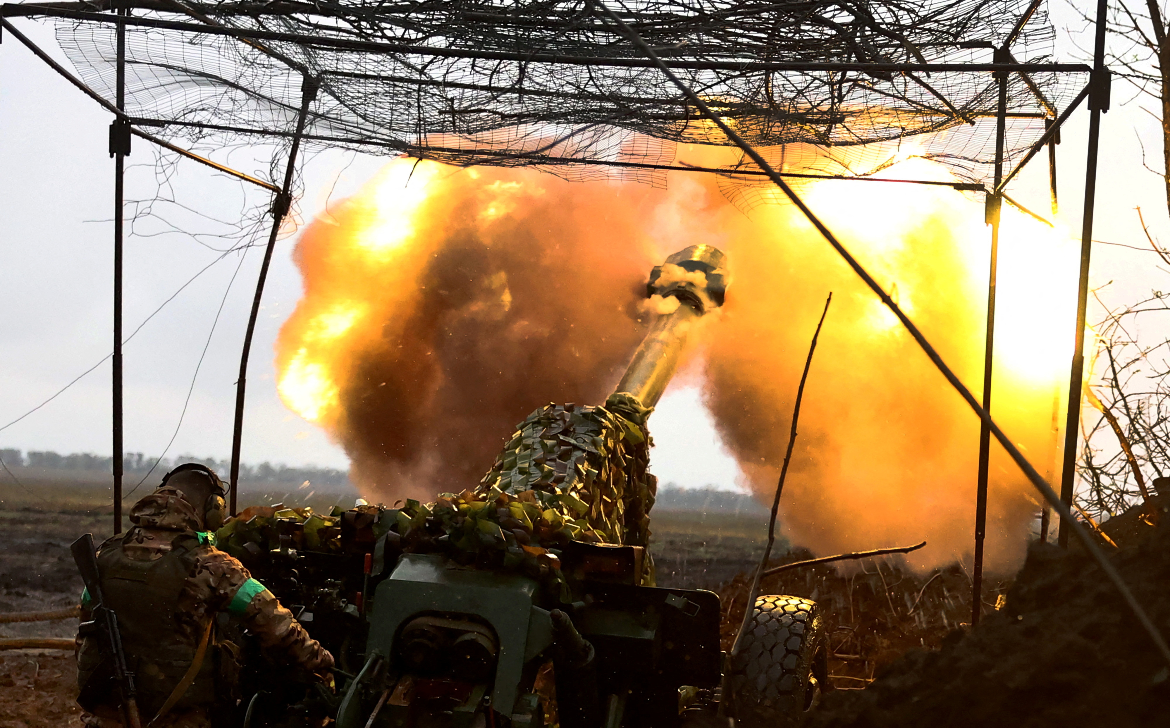 Ukrainian artillery fires towards the frontline near Bakhmut. The gun is hidden beneath camouflage. There is a smoke and orange flame.
