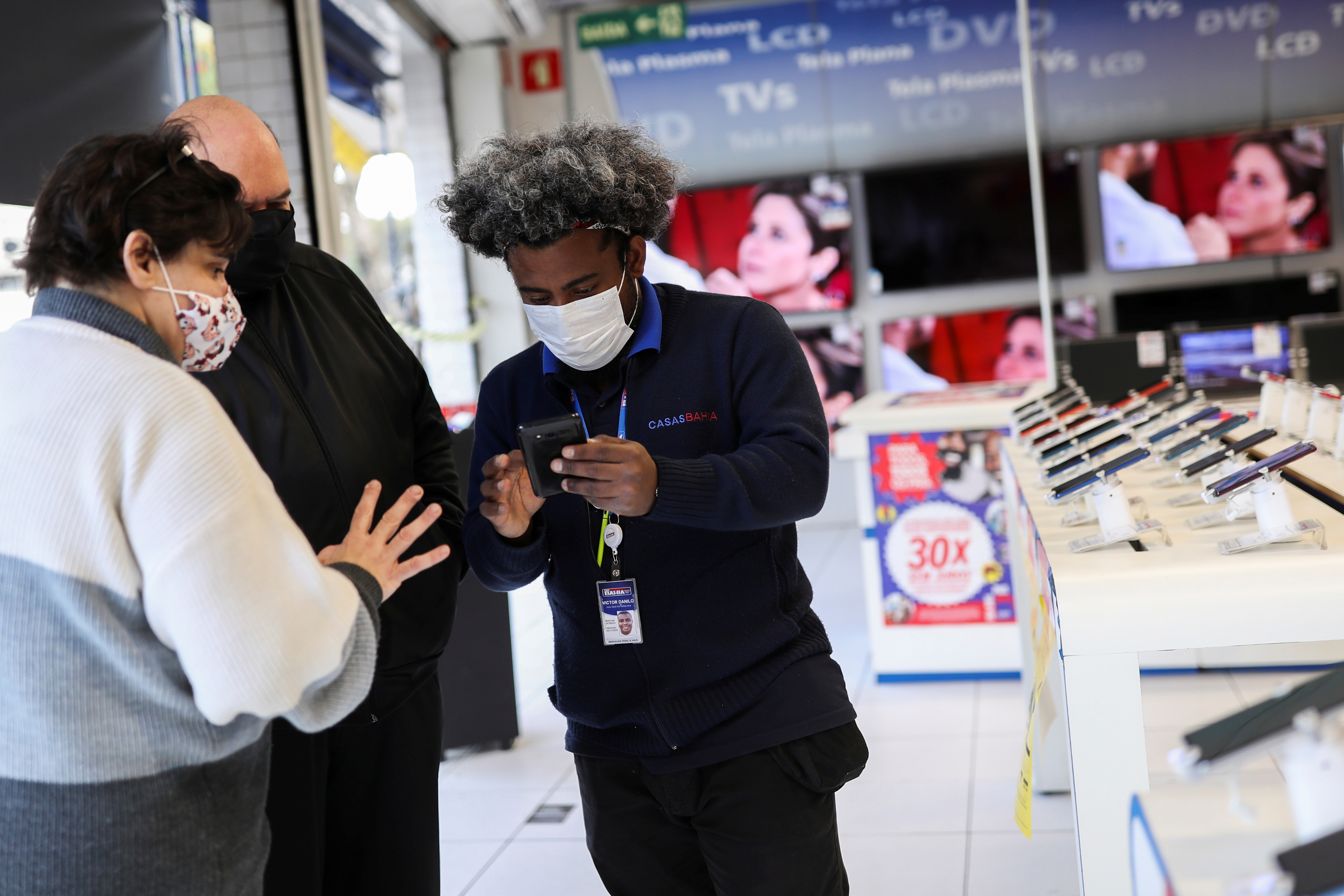 Vendor Victor Danilo dos Santos Silva holds a phone as he shows mobile phones on sale on the store website to customers at a Casas Bahia store in Sao Paulo, Brazil