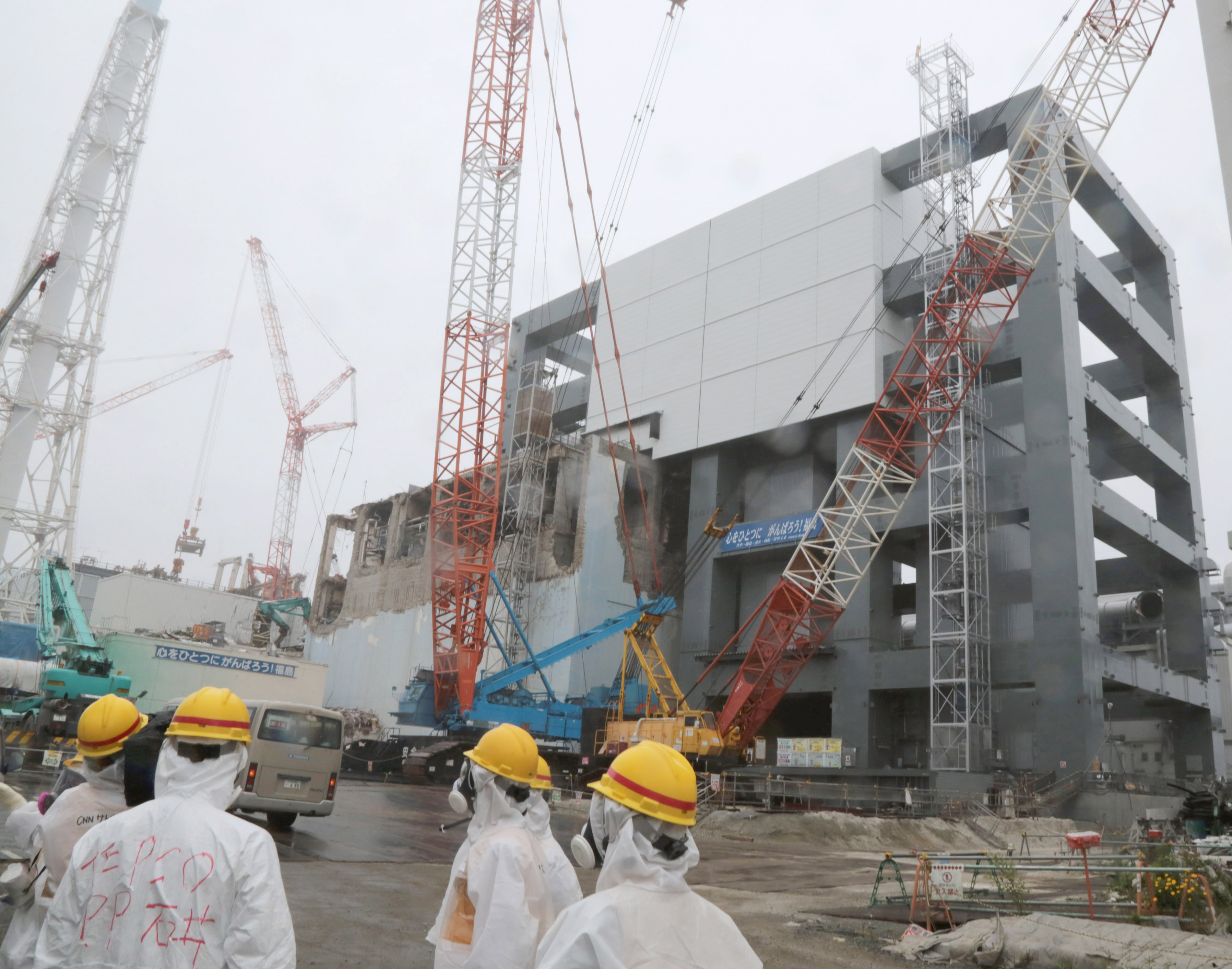 Workers monitoring the damaged plant in 2013, They are outside and wearing full hazmat suits and helmets. There are cranes around the building.