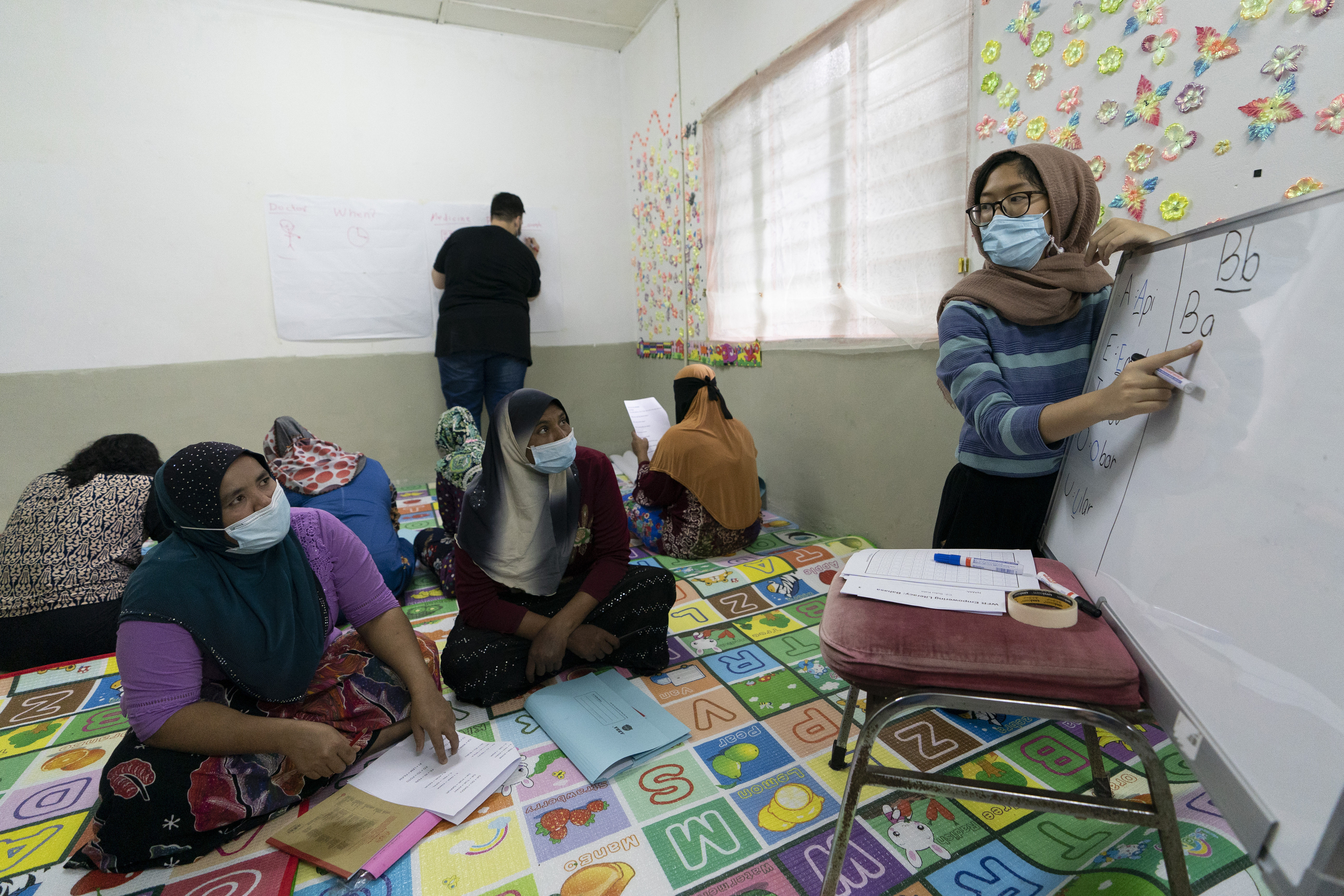 Refugee women in an English lesson at a community school run by volunteers. They are seated on the floor and their teacher is pointing to letters on a white board. More women are seated behind them facing the opposite wall and having lesson from a different teacher.