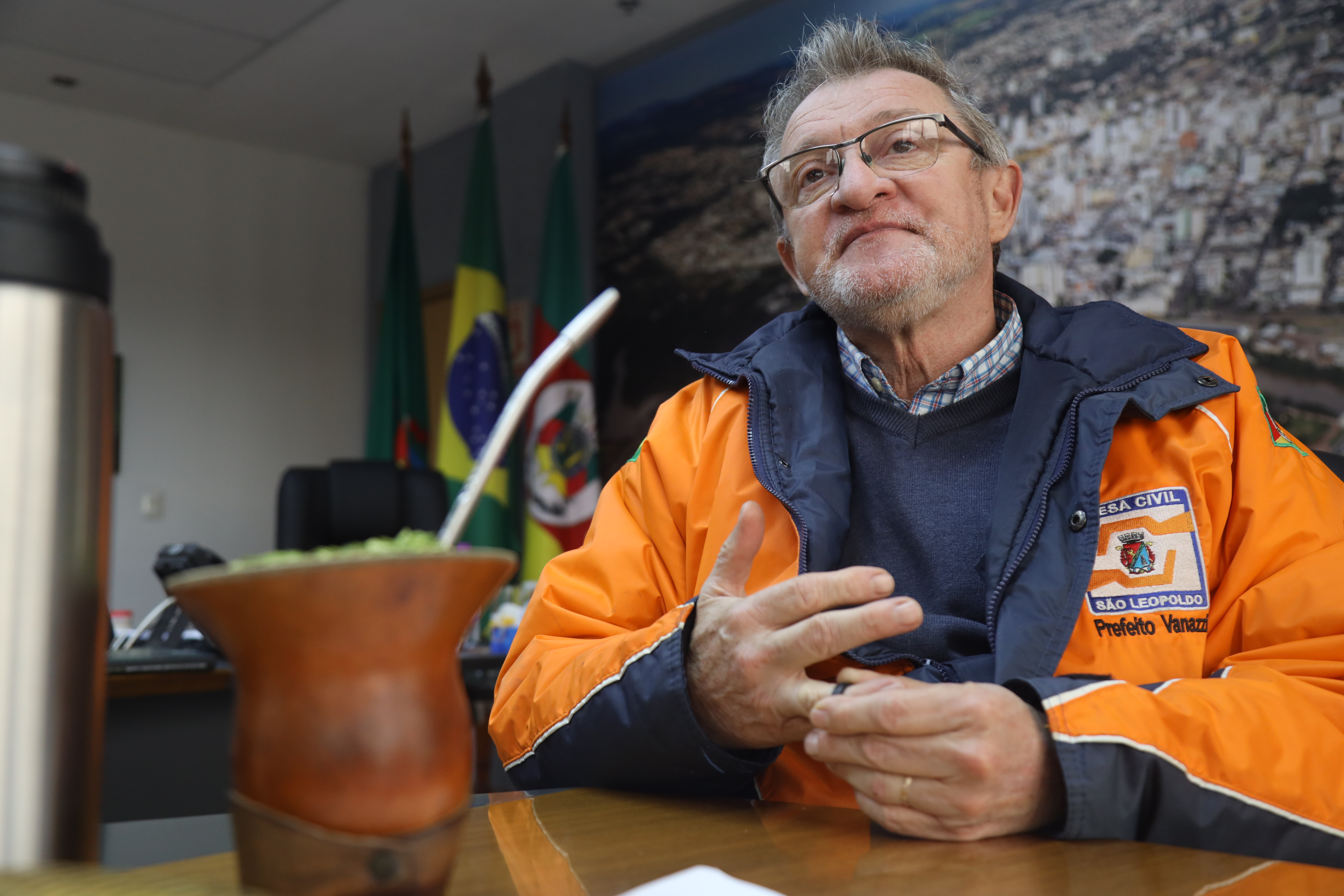 Ary Vanazzi, the mayor of São Leopoldo, sits at a table in an orange and blue windbreaker jacket.