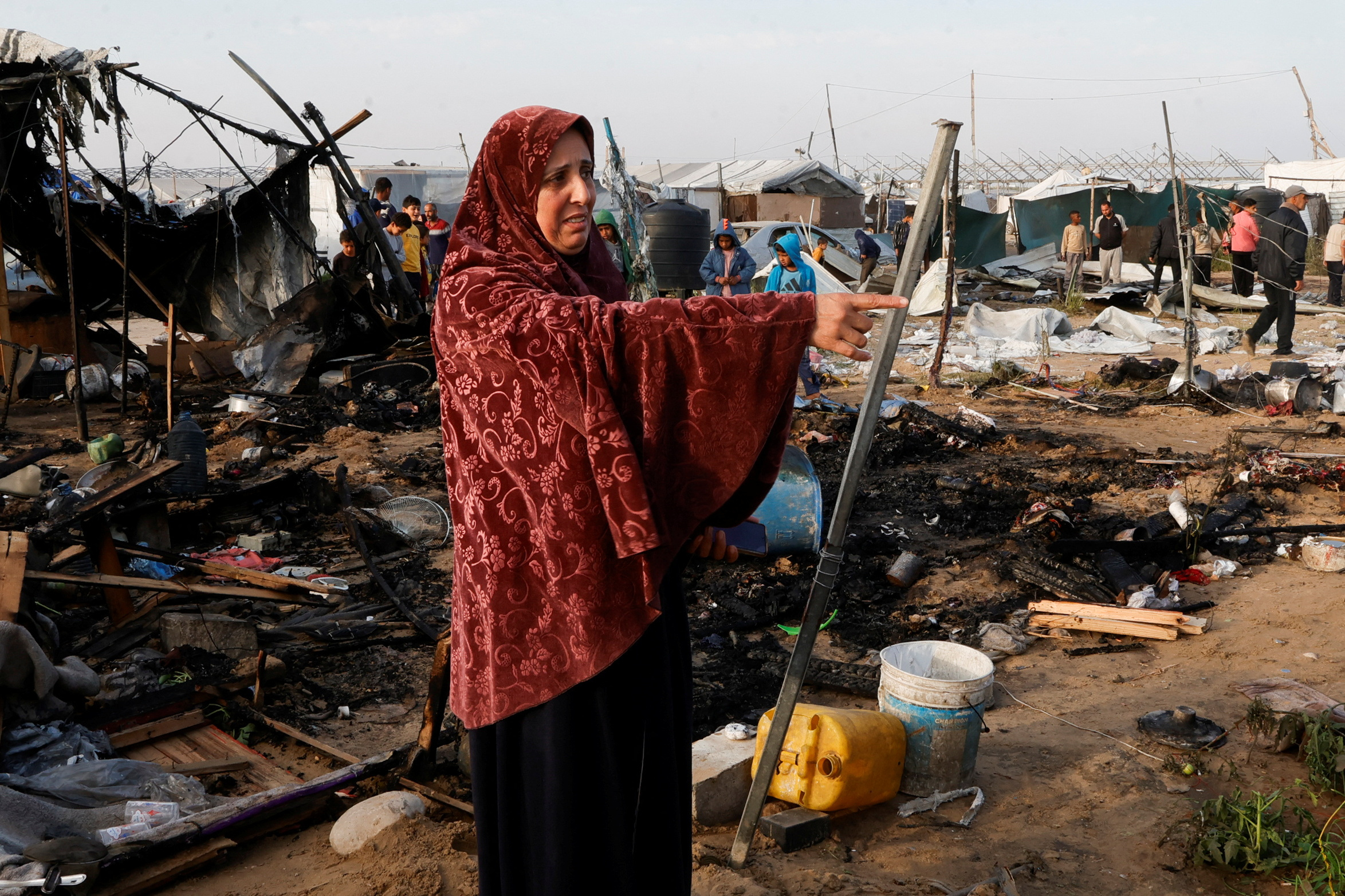 a woman wearing a red head scarf gestures near the charred remains of a tent