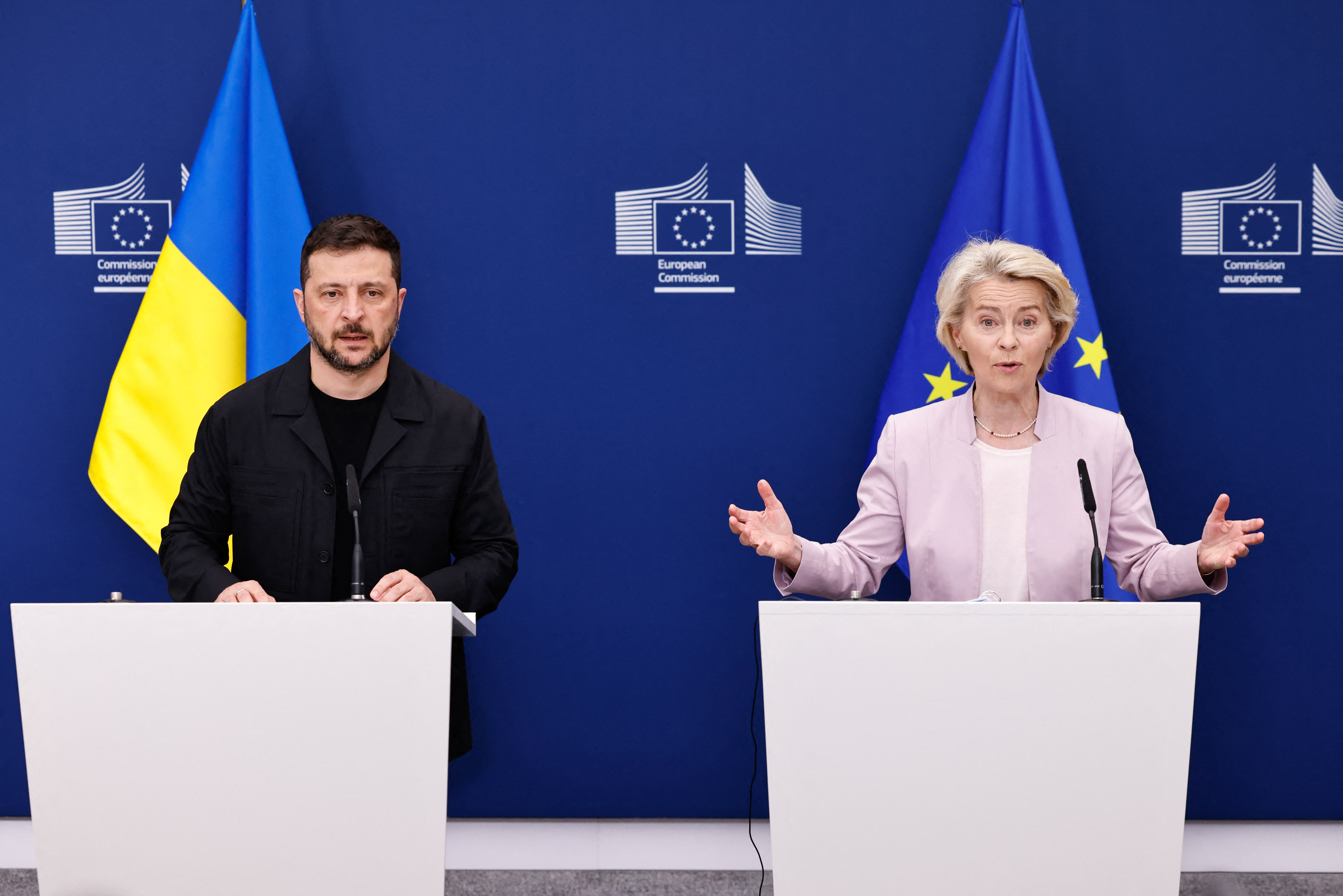 a man wearing all black and a woman in a light pink suit make speeches in front of the Ukrainian and EU flags