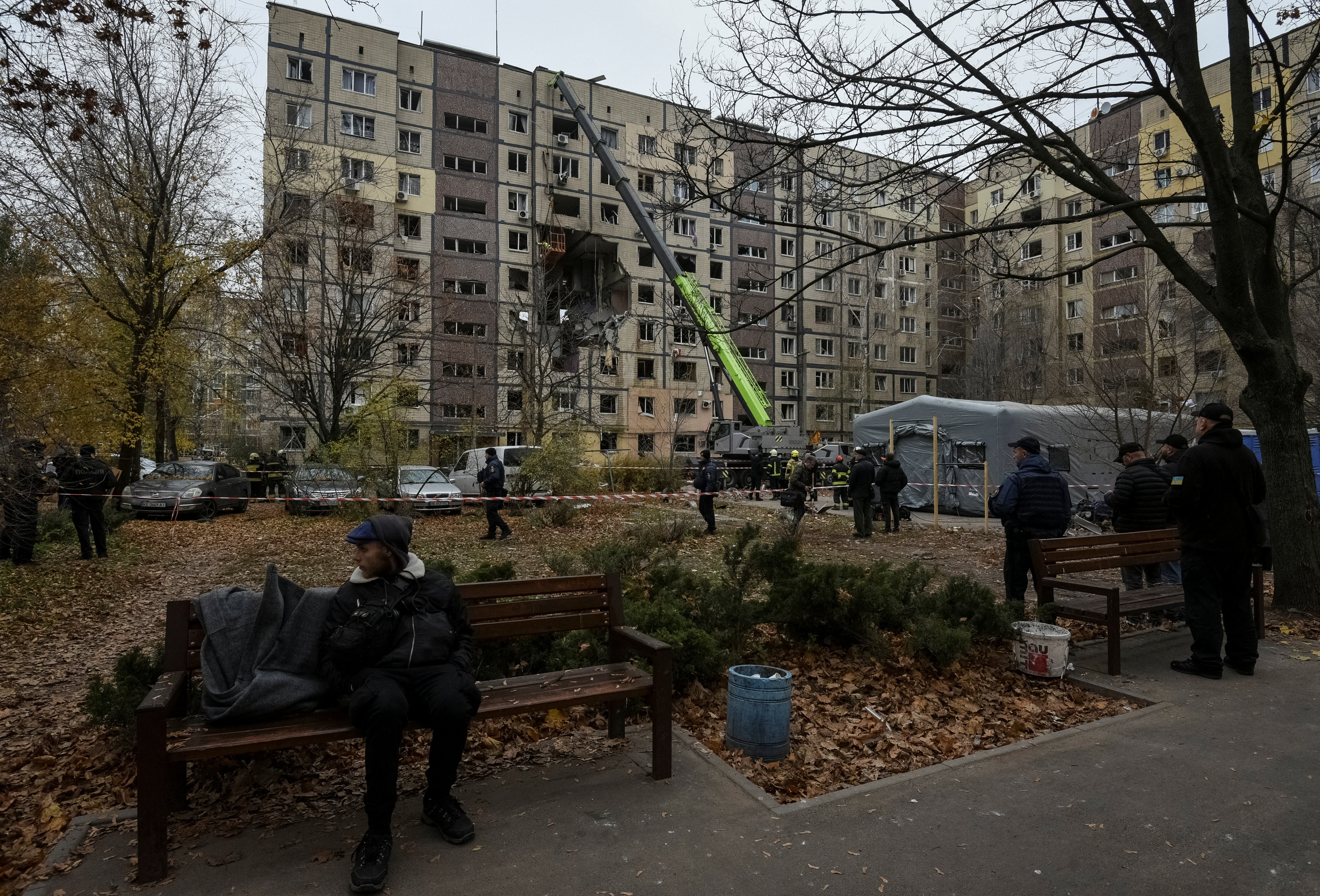 A man sits on a bench as rescuers and others stand near damaged apartment buildings.