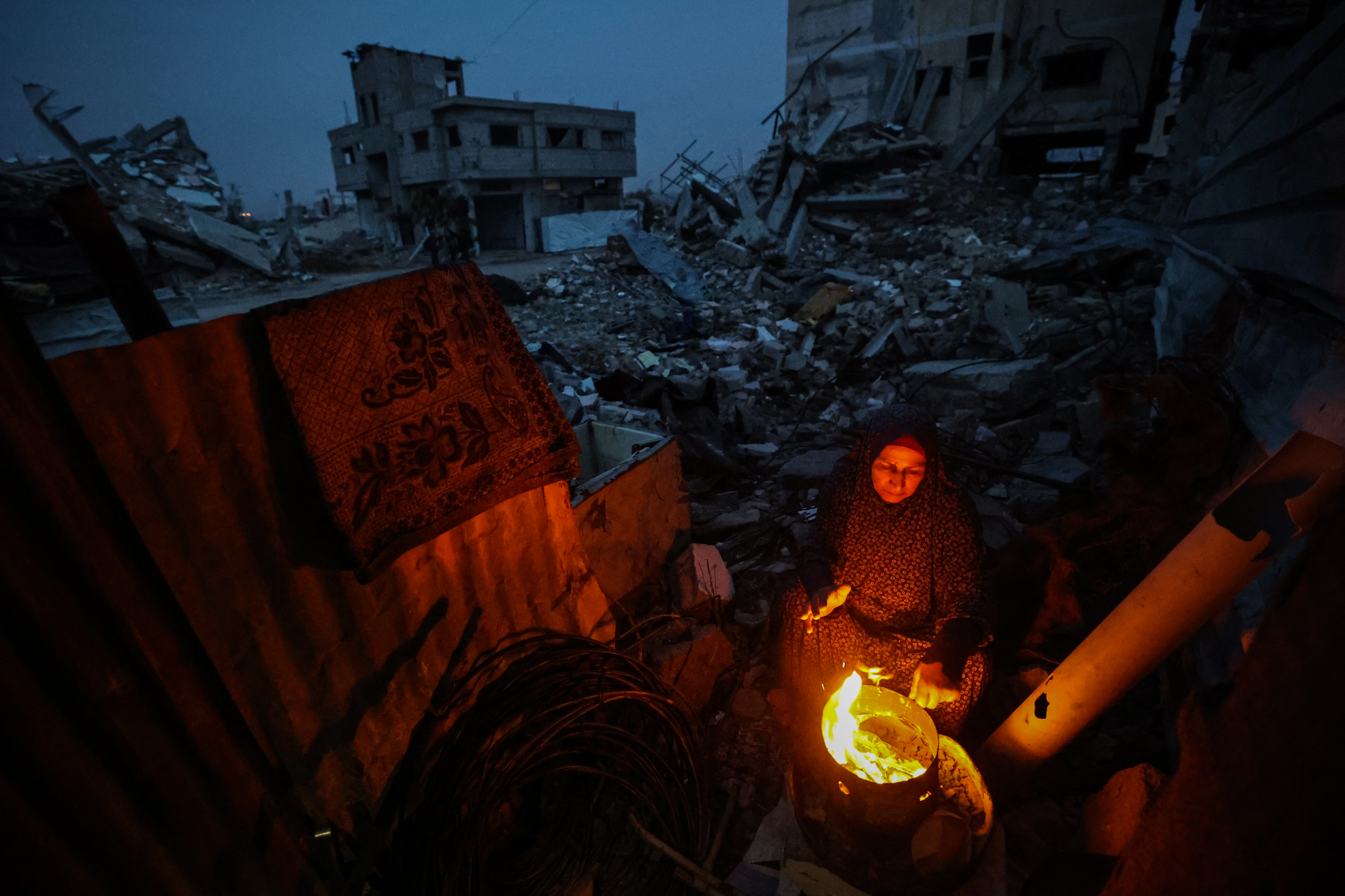 A displaced Palestinian woman warms herself beside a fire amid the ruins of destroyed buildings in the Bureij refugee camp, in the central Gaza Strip