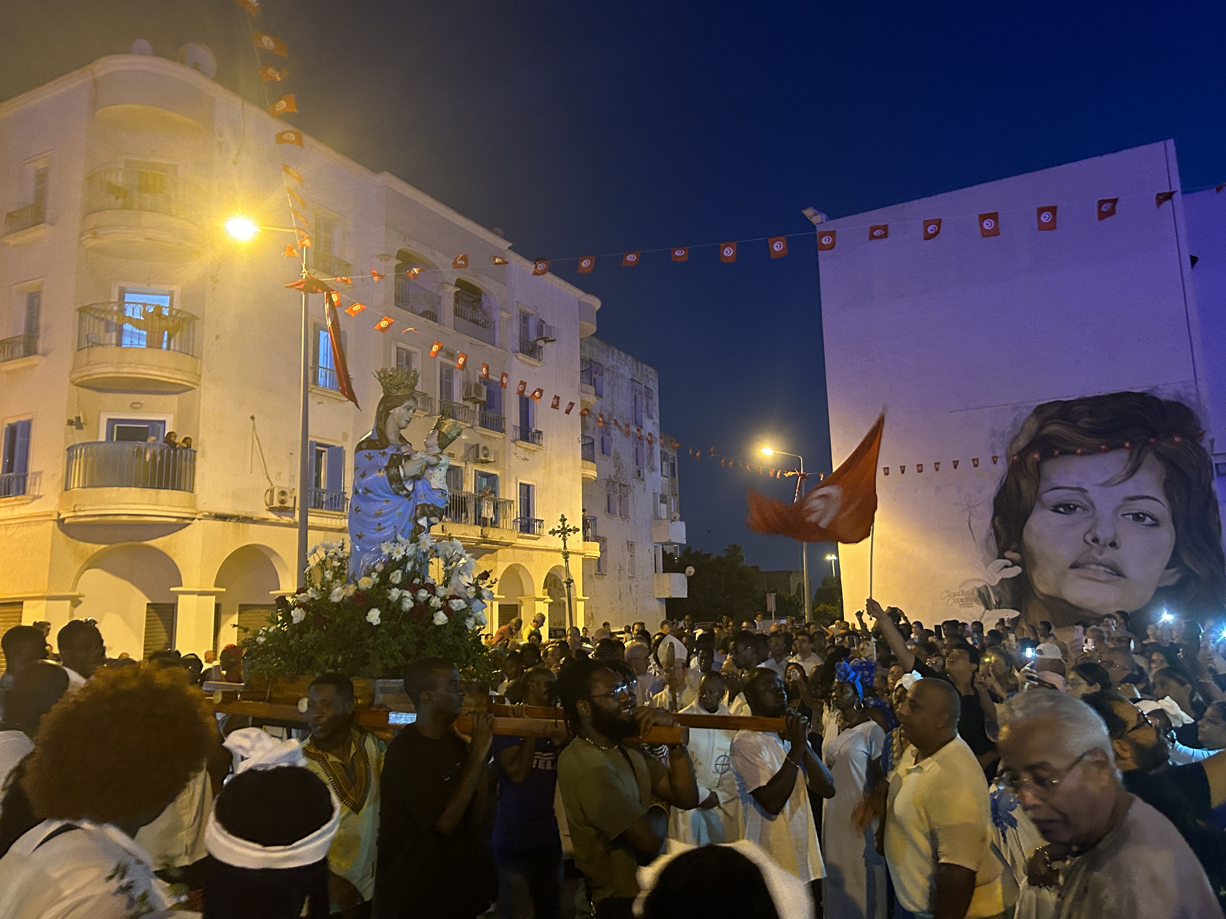 A crowd carry a statue of the Virgin Mary in a square, with a mural depicting Claudia Cardinale on a wall