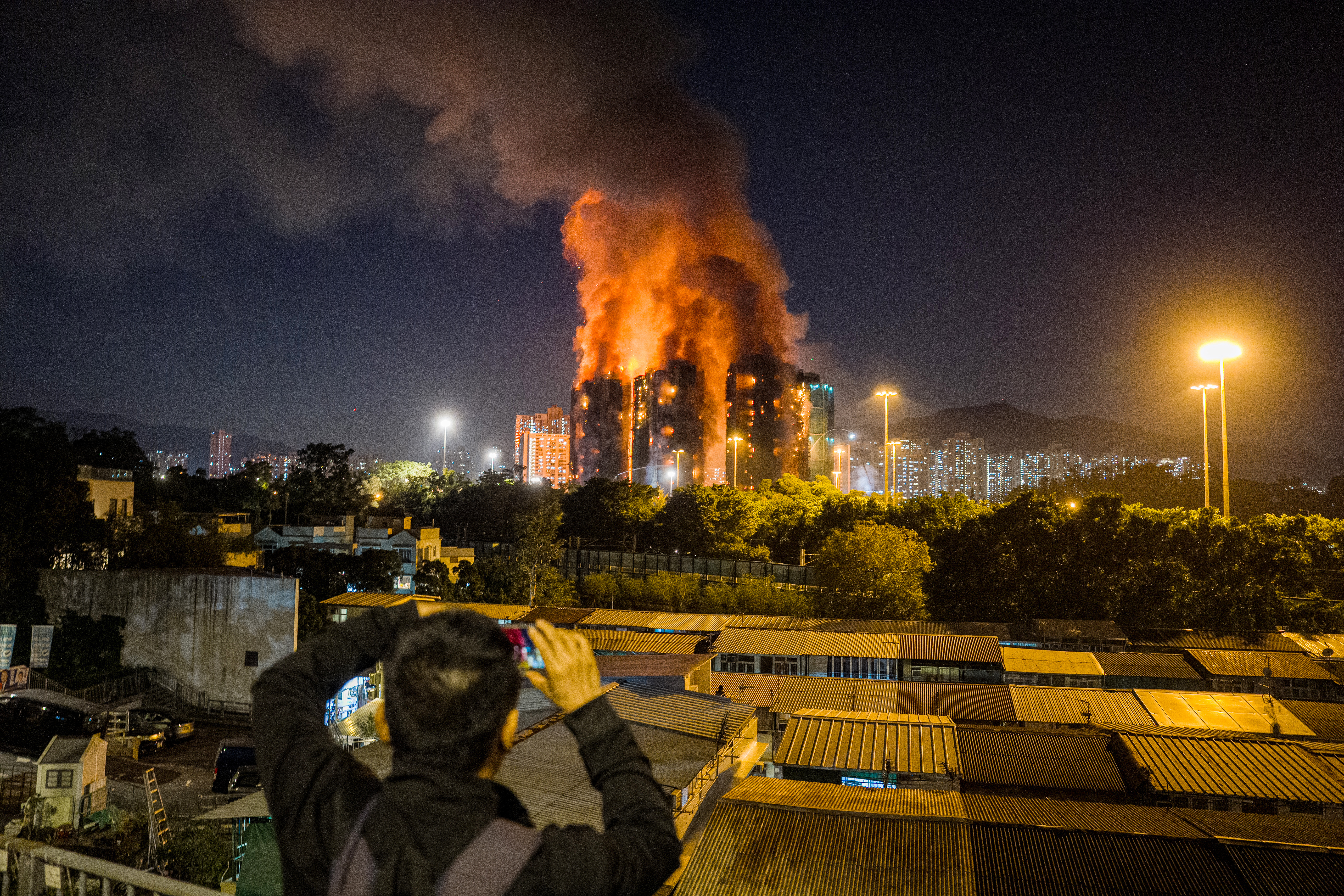 An onlooker takes photos as a major fire engulfs several apartment blocks at the Wang Fuk Court residential estate (background) in Hong Kong's Tai Po district on November 26, 2025.