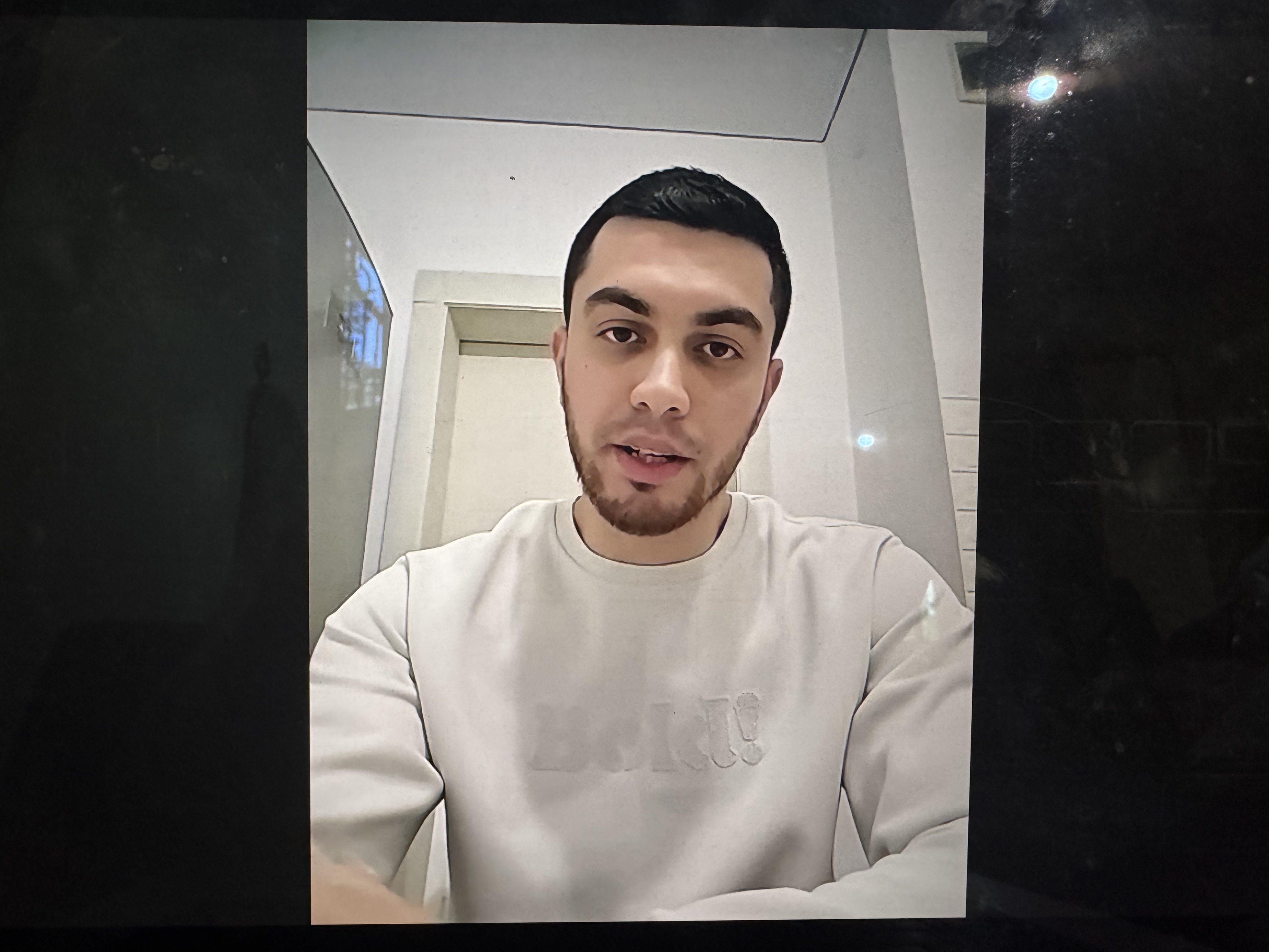 A photo of a computer screen with the image of Ilyas Abu Safiya on a video call. A clean-shaven young man with dark hair. Reflected in the computer screen is a streetlight because the journalists could only get enough internet to run an online interview by standing in the street, due to Israel's blockade of all services and goods in Gaza