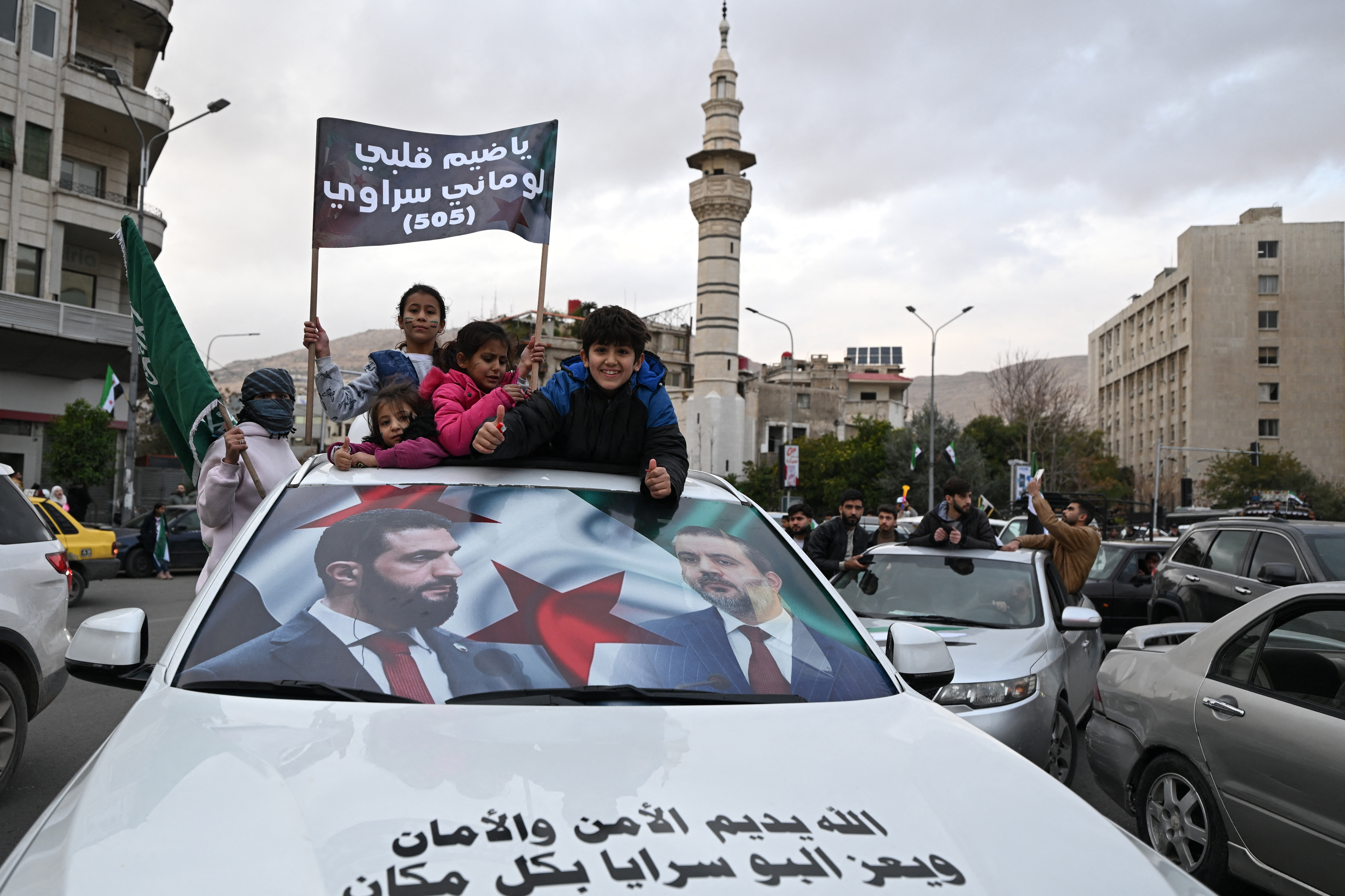 People ride a car reflecting the faces of Syria's President Ahmed al-Sharaa and Syrian Foreign Minister Asaad al-Shaibani as they celebrate a year since the ousting of longtime ruler Bashar al-Assad in Damascus