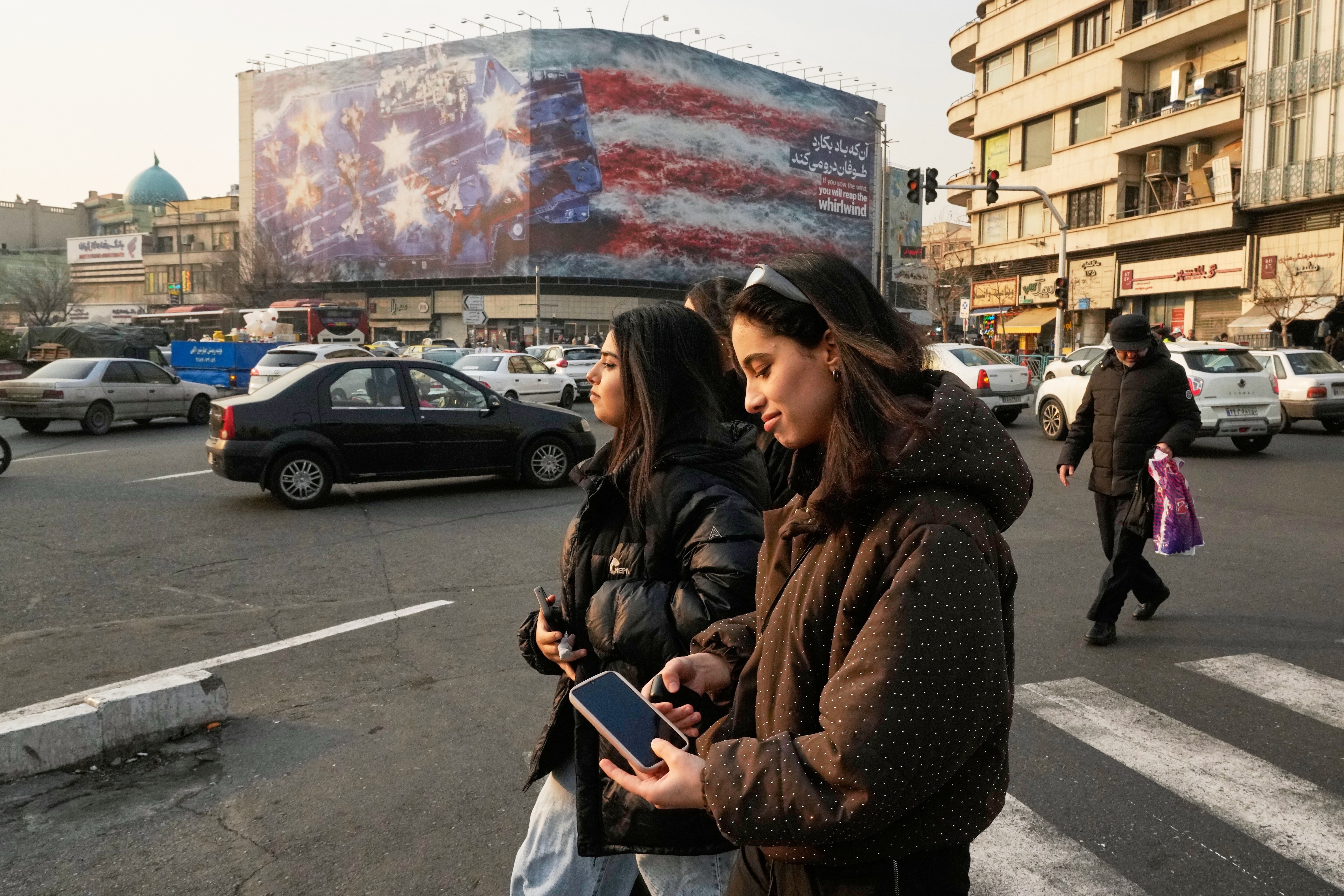 Two girls, not wearing the legally required headscarves, walk past a billboard depicting a damaged U.S. aircraft carrier with disabled fighter jets on its deck and a sign reading in Farsi and English, "If you sow the wind, you'll reap the whirlwind," at Enqelab-e-Eslami (Islamic Revolution) Square in Tehran, Iran, Sunday, Jan. 25, 2026. (AP Photo/Vahid Salemi)