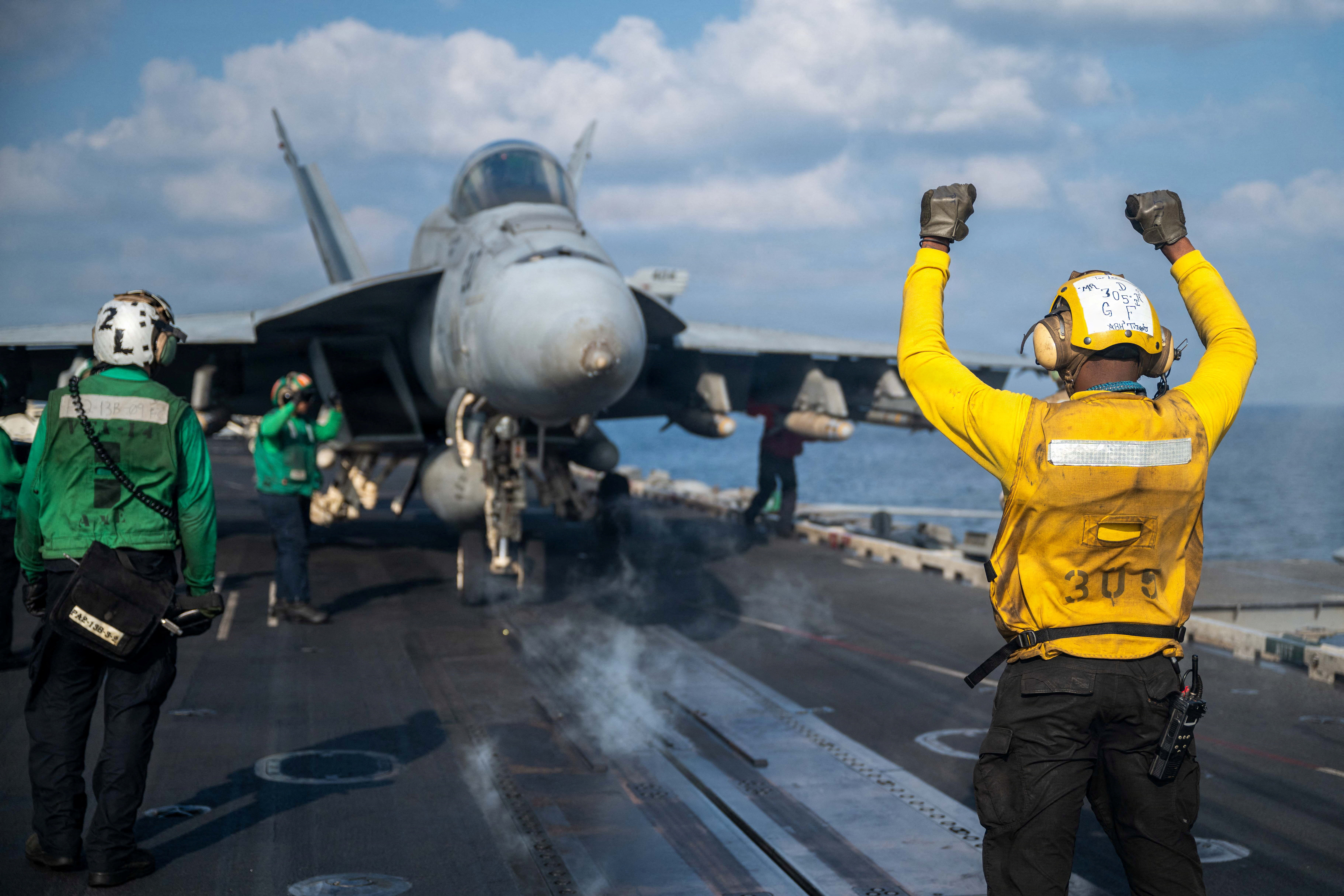 Man in yellow stands and signals in front of fighter jets.