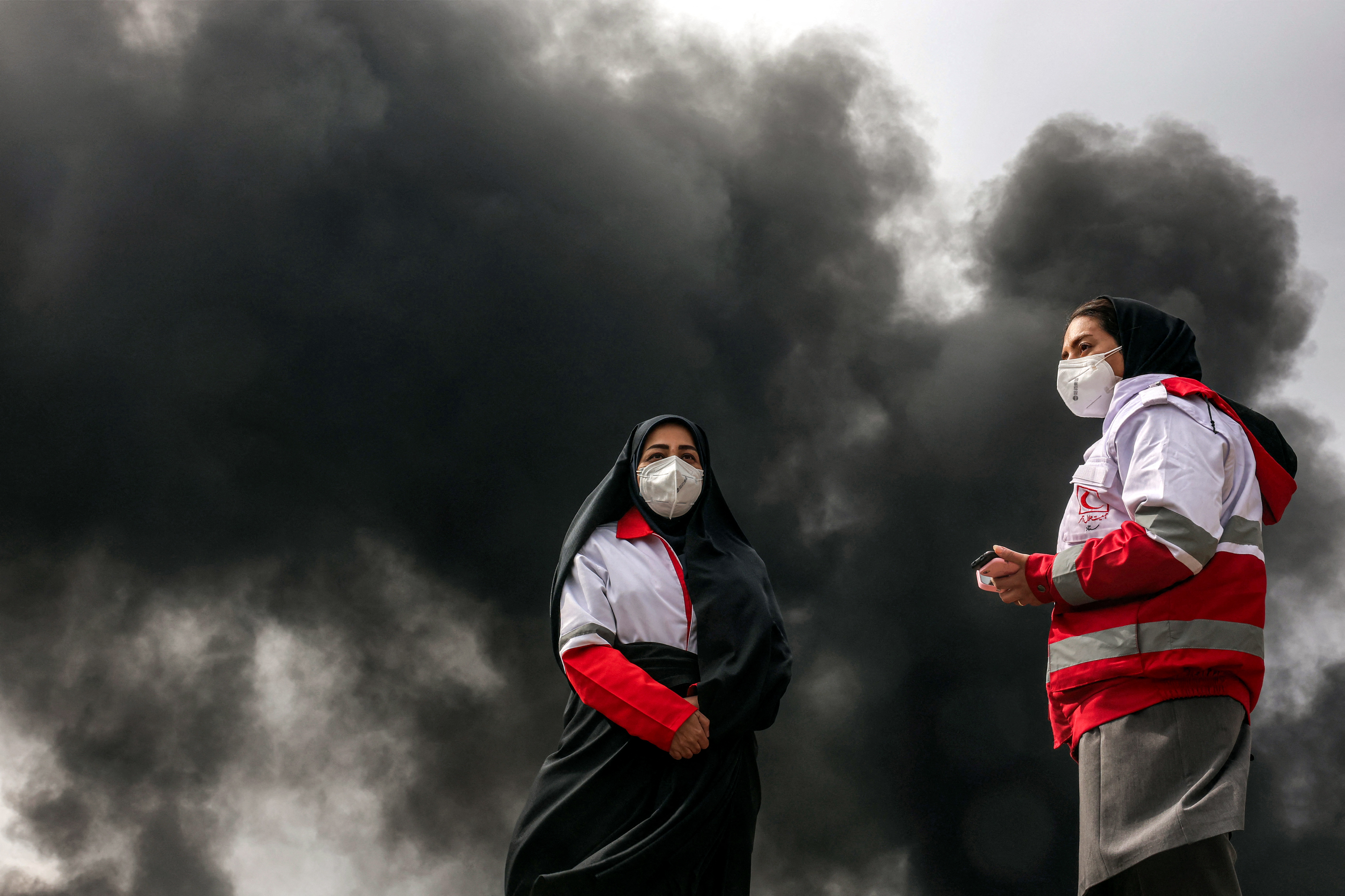 Women members of Iran's Red Crescent society stand near smoke plumes from an ongoing fire following an overnight airstrike on the Shahran oil refinery in northwestern Tehran on March 8, 2026.