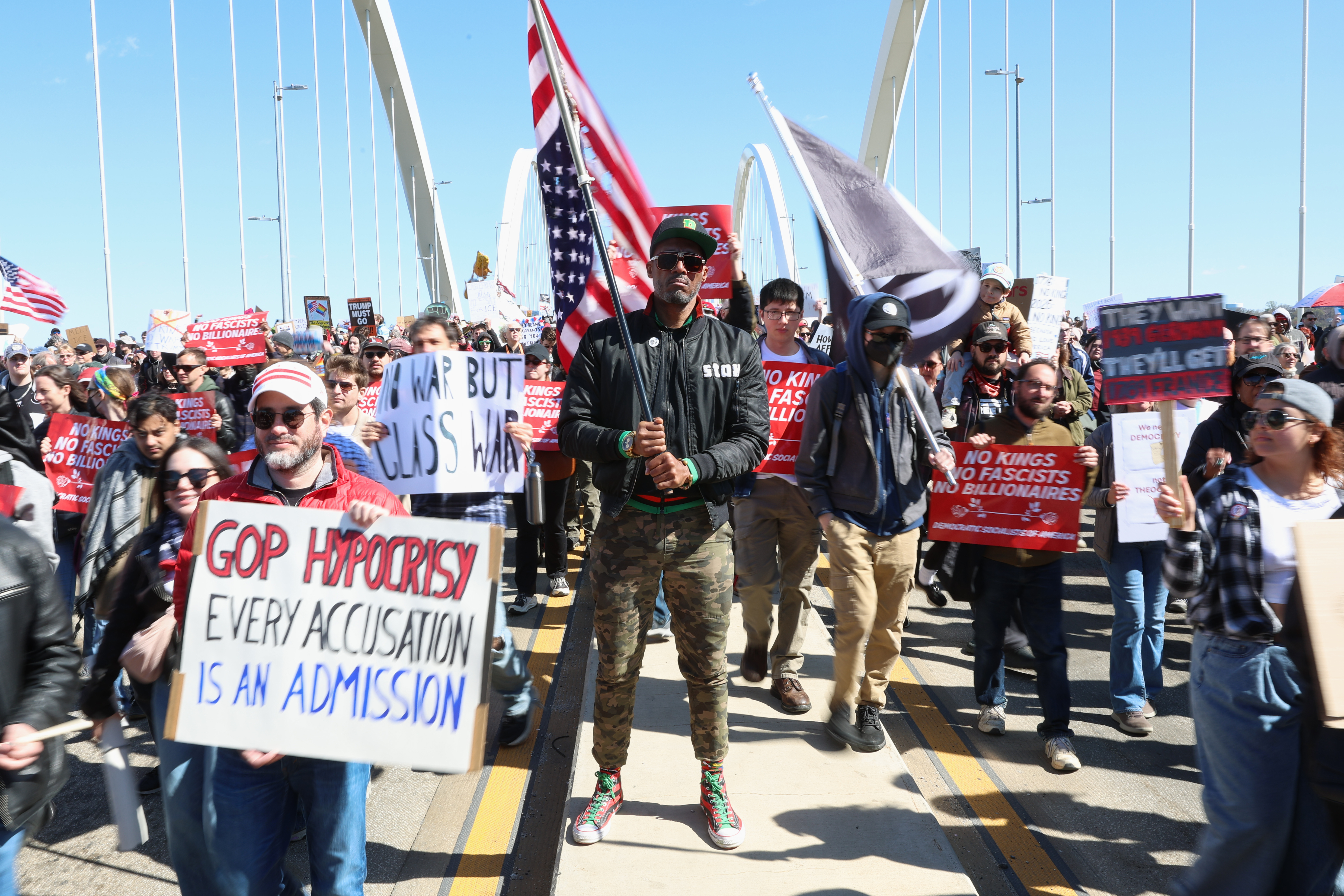 Kymone Freeman holds an American flag while demonstrators stream past him over the Frederick Douglass Bridge during the No Kings protest in Washington, Saturday, March 28, 2026. (AP Photo/Tom Brenner)