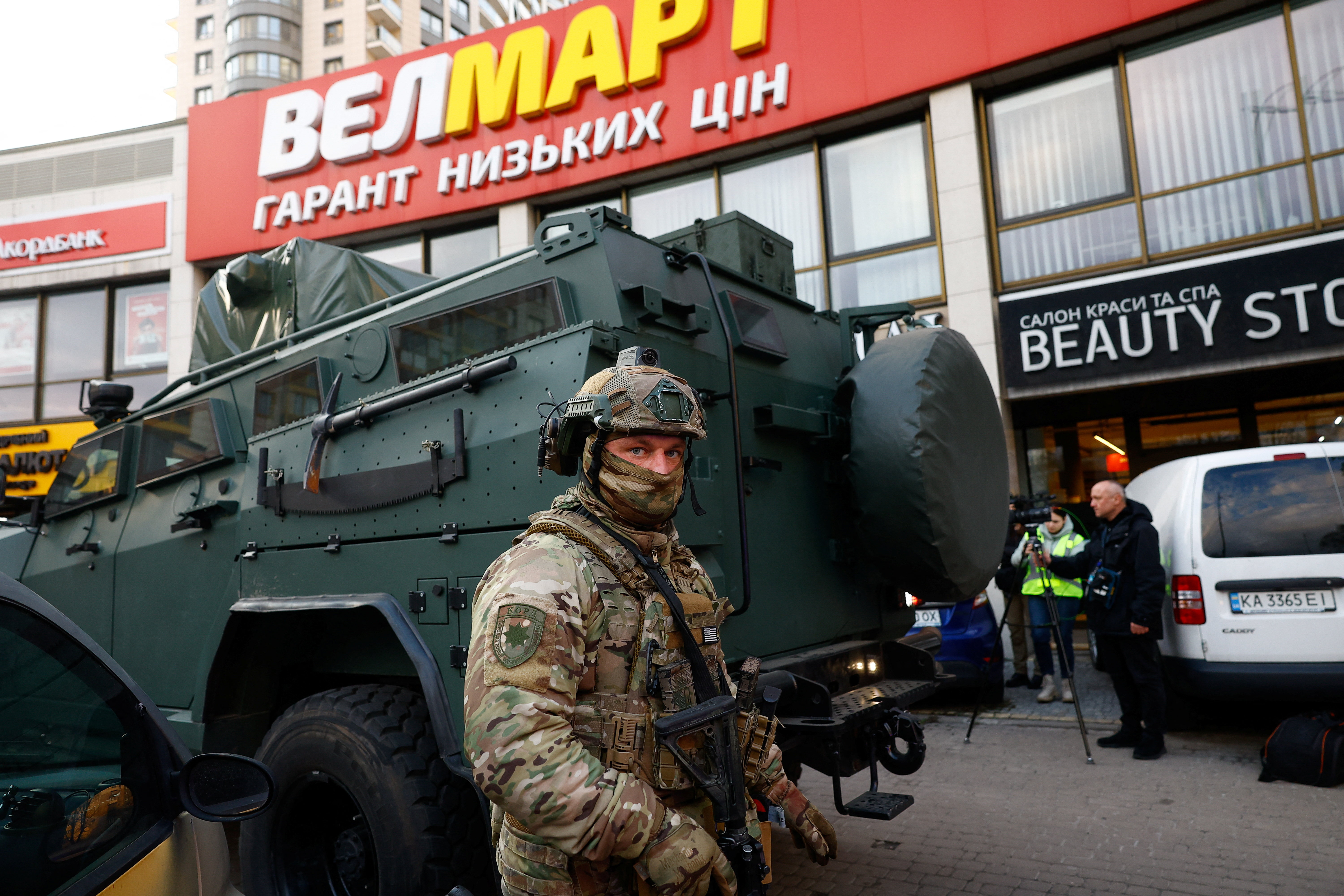 A service member stands by an armoured vehicle outside a supermarket.