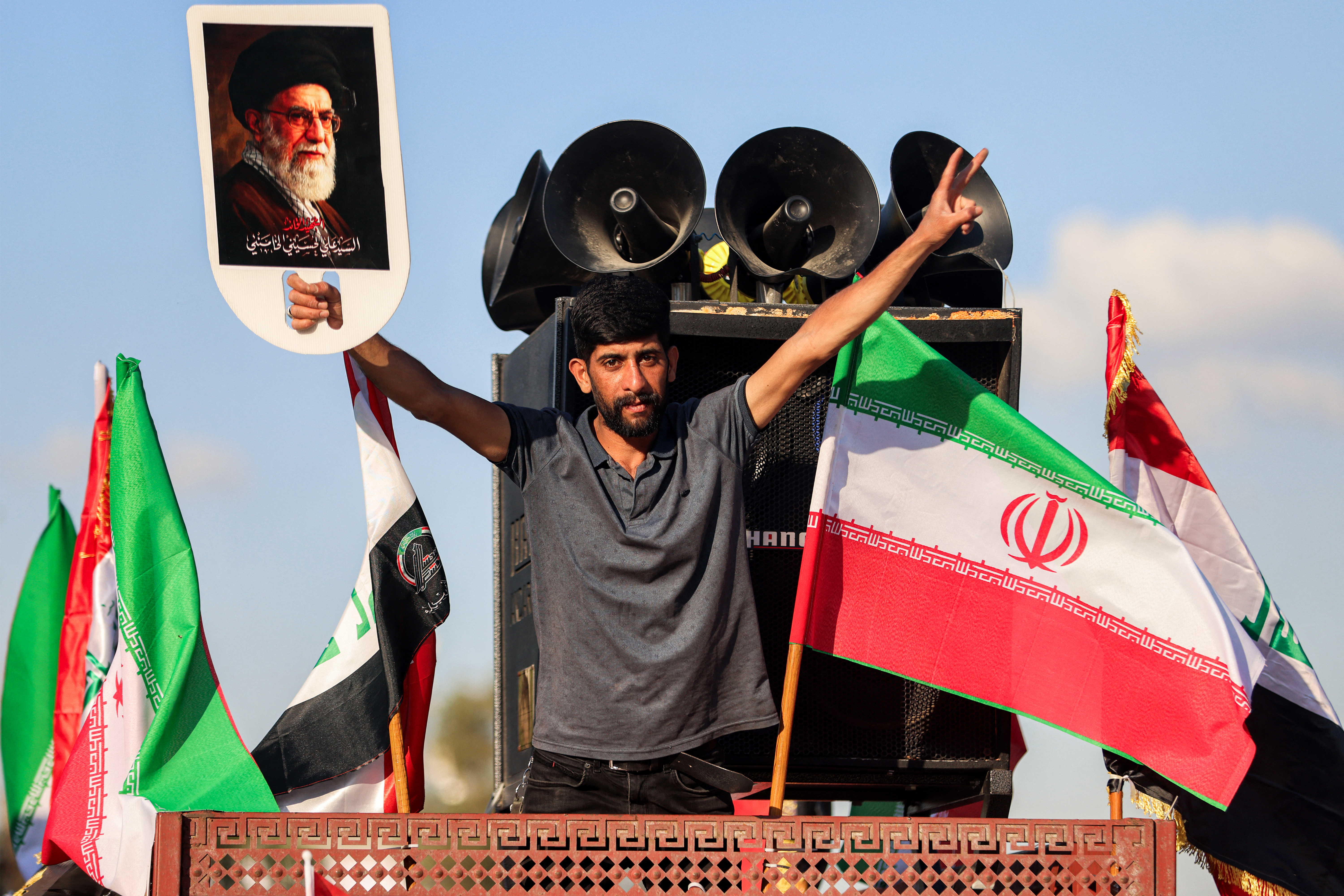 A man gestures with picture of Iran's slain supreme leader Ayatollah Ali Khamenei next to Iranian and Iraqi flags from a atop a truck during celebrations welcoming the two-week ceasefire between the United States and Iran in Baghdad's central Tahrir Square on April 8, 2026. Iraqi armed factions loyal to Iran and operating under the banner of the "Islamic Resistance in Iraq" announced a two-week halt to attacks they had launched since the start of the war in the Middle East against "enemy bases" in Iraq and the region. The announcement came shortly after Washington and Tehran declared a ceasefire for the same period. [AHMAD AL-RUBAYE / Reuters]