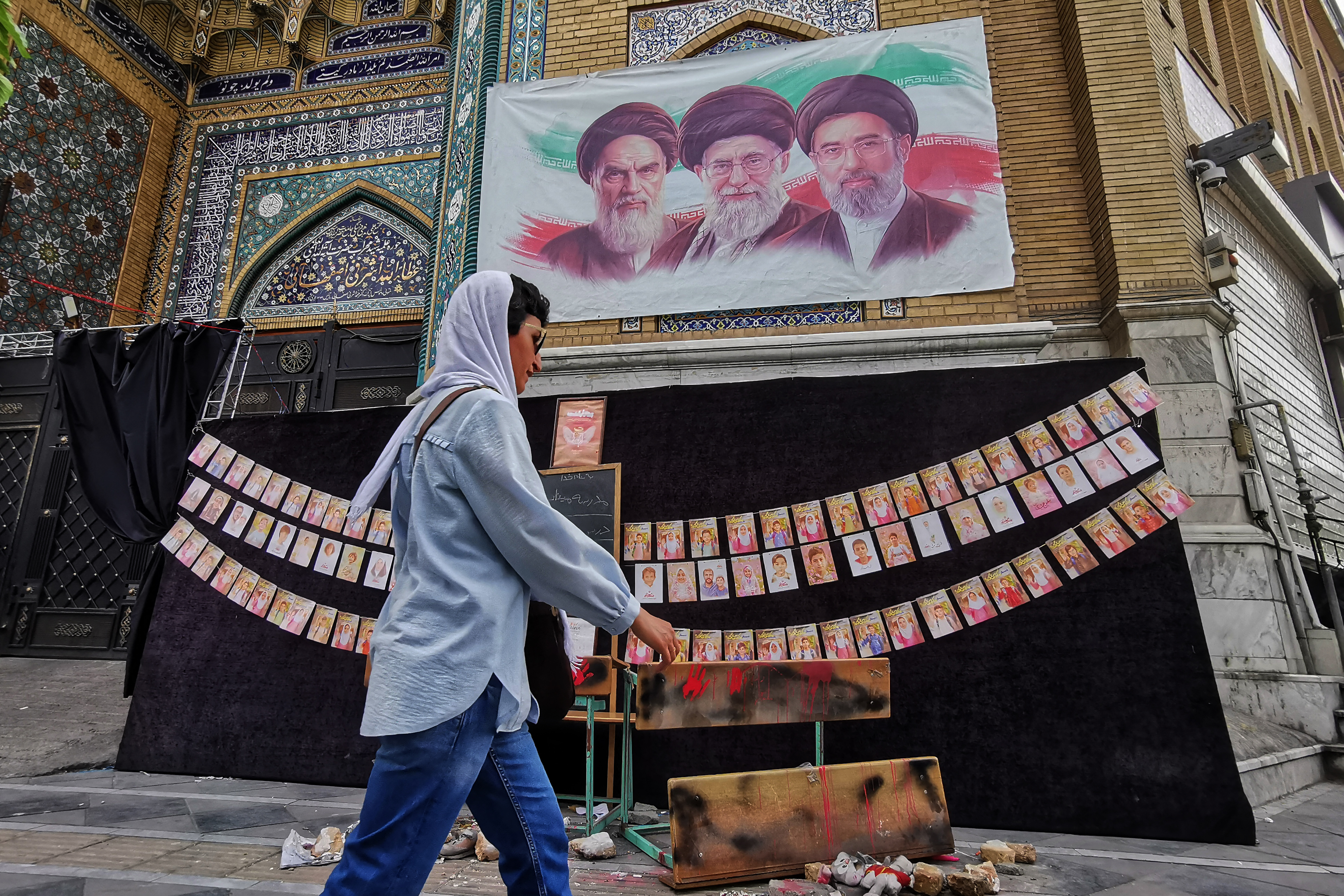 A woman walks past images of Iran's late supreme leaders Ayatollah Ruhollah Khomeini (above L) and Ayatollah Ali Khamenei (Above C) next to newly elected supreme leader Mojtaba Khamenei (above R), and photos strung along the wall of children killed on the first day of the war in an alleged US-Israeli missile strike on a school in the southern Iranian city of Minab, outside a mosque in the capital Tehran on April 25, 2026. On February 28, Israel and the United States launched strikes on Iran, killing its supreme leader and triggering a war that spread across the Middle East.