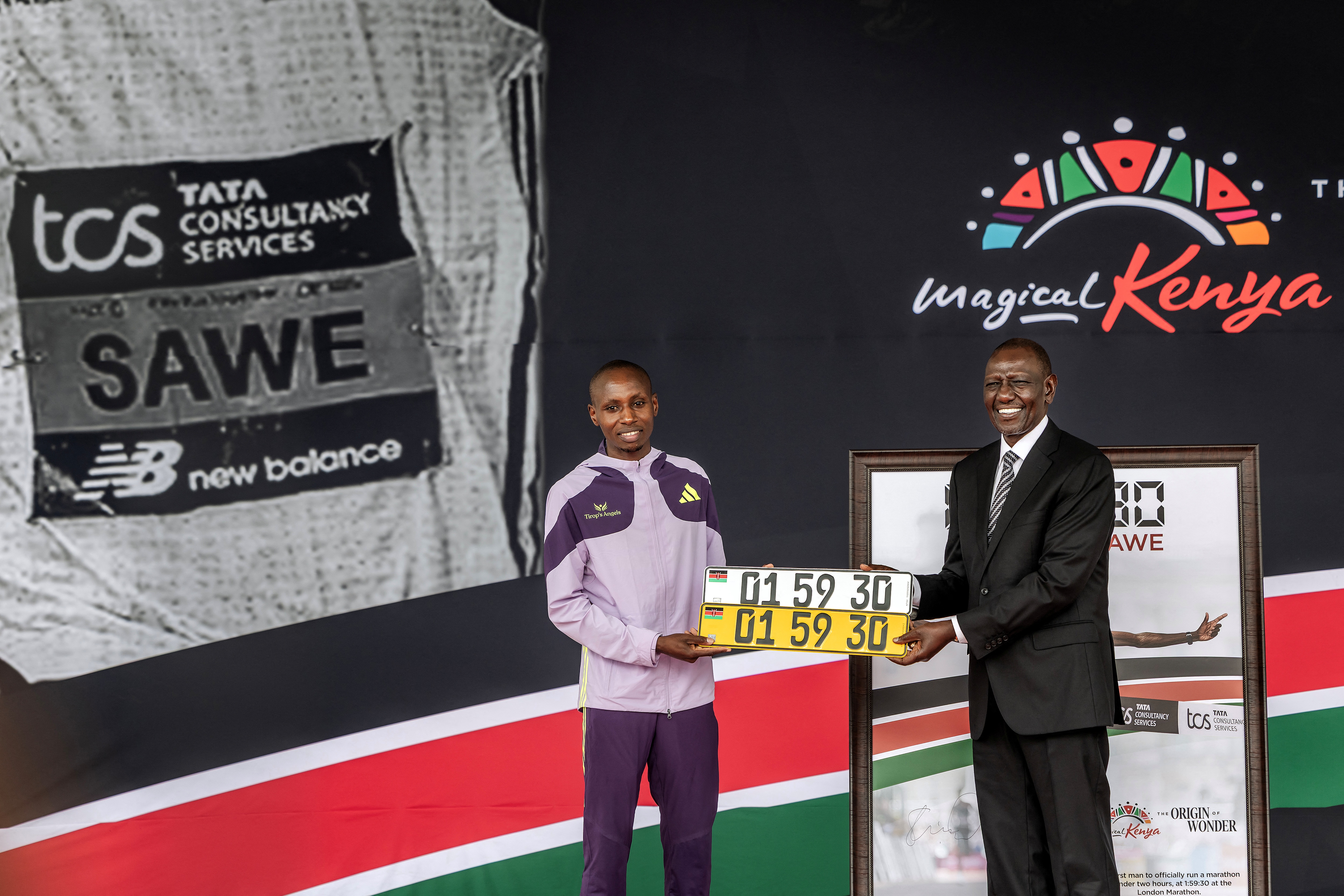 Kenya's President William Ruto (R) gifts a plaque reading Kenyan marathon runner Sabastian Sawe (L) record time after winning the London Marathon as he congratulates the athlete on on stage, during a ceremony at State House in Nairobi, on April 30, 2026. Sabastian Sawe became the first man to run under two hours for a marathon in race conditions thanks to extraordinary talent, a punishing training regime, and the lightest shoes in history. In London on April 26, 2026, the 31-year-old Kenyan achieved one of sport's milestones, ranking alongside Roger Bannister's first sub-four minute mile in 1954. (Photo by Luis TATO / AFP)