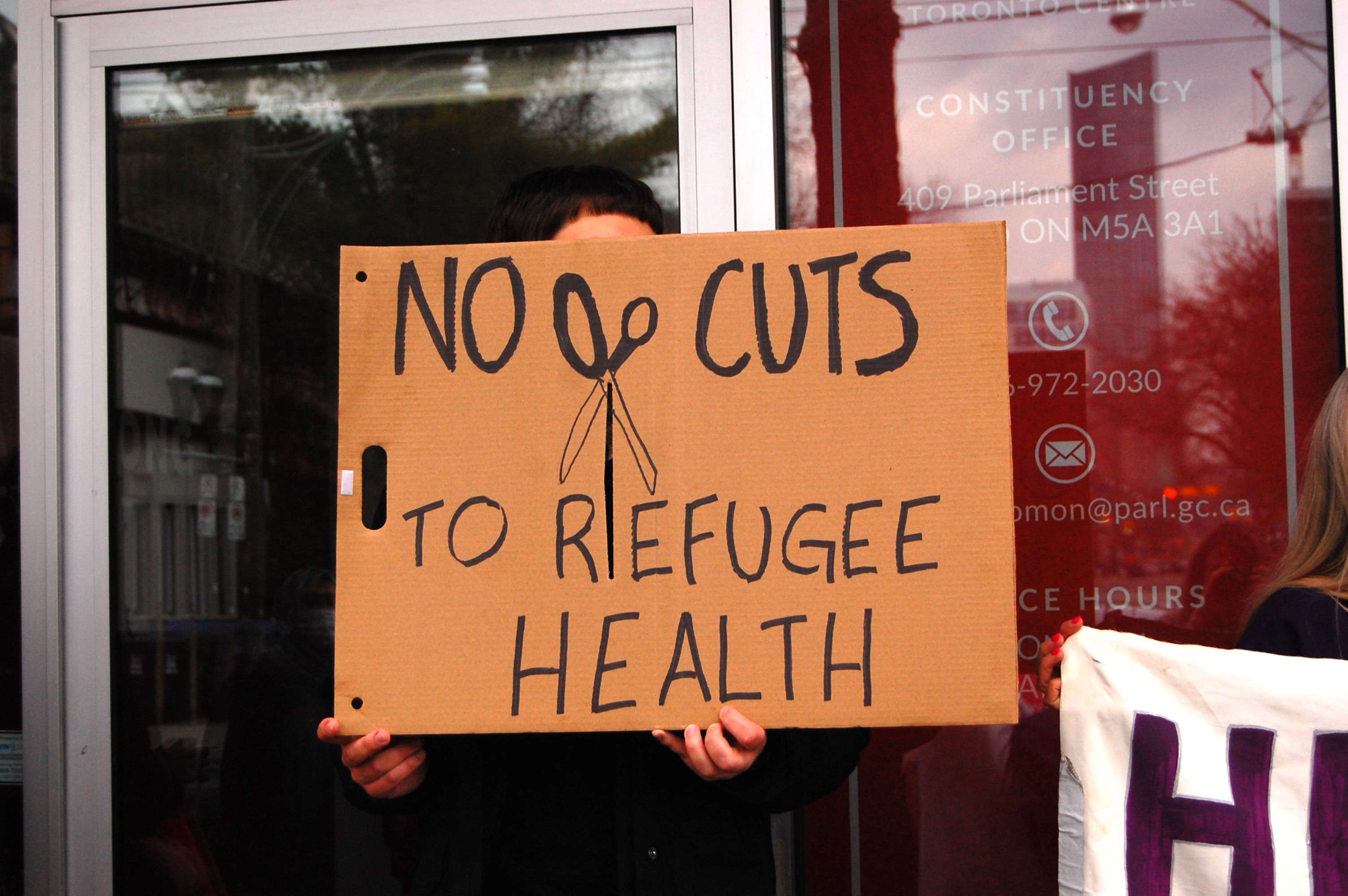 A protester holds a sign that reads, ‘No cuts to refugee health’ at the rally in Toronto, Canada, April 14, 2026
