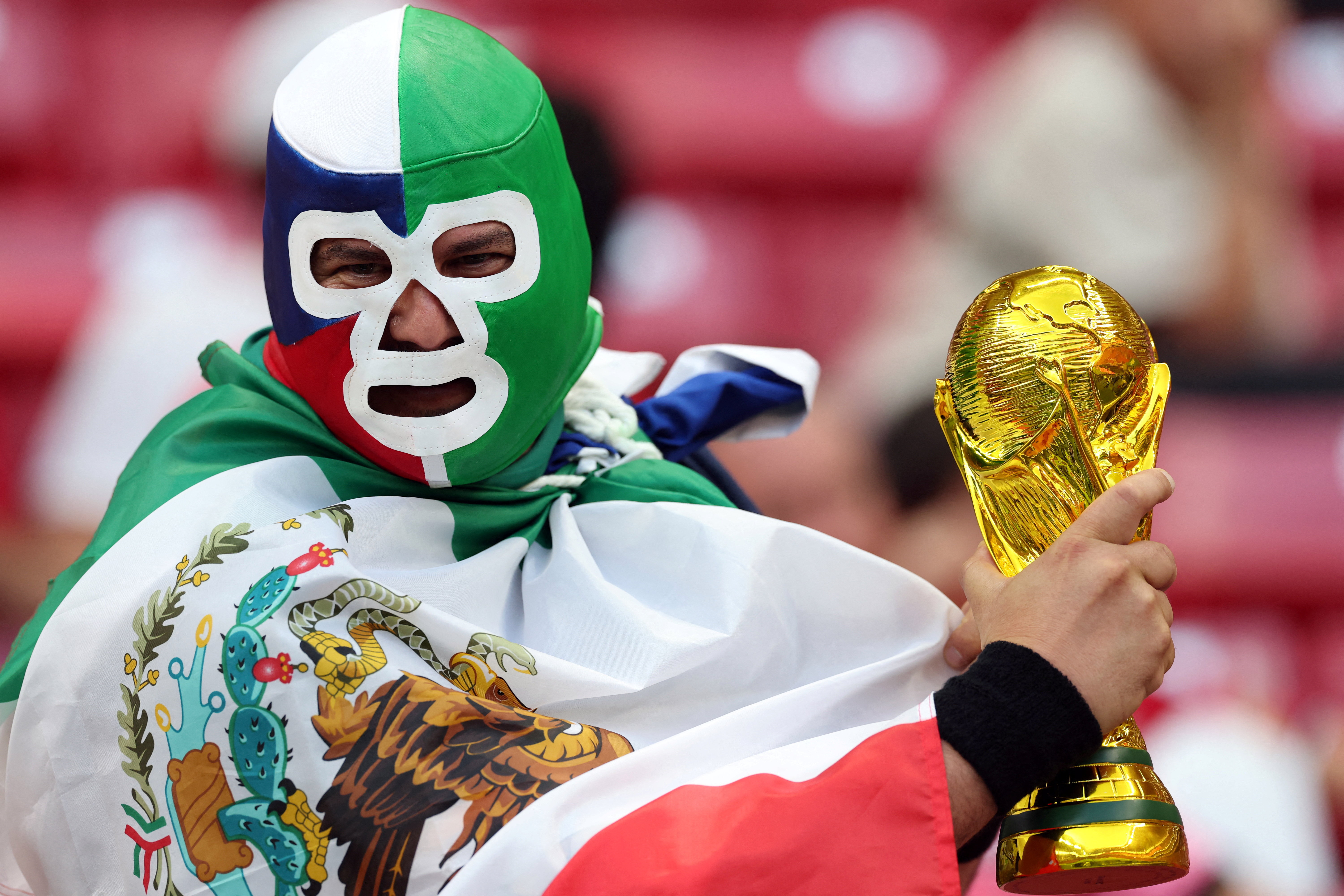A Mexico fan wearing a wrestling mask holds a replica world cup trophy in the stands before the match