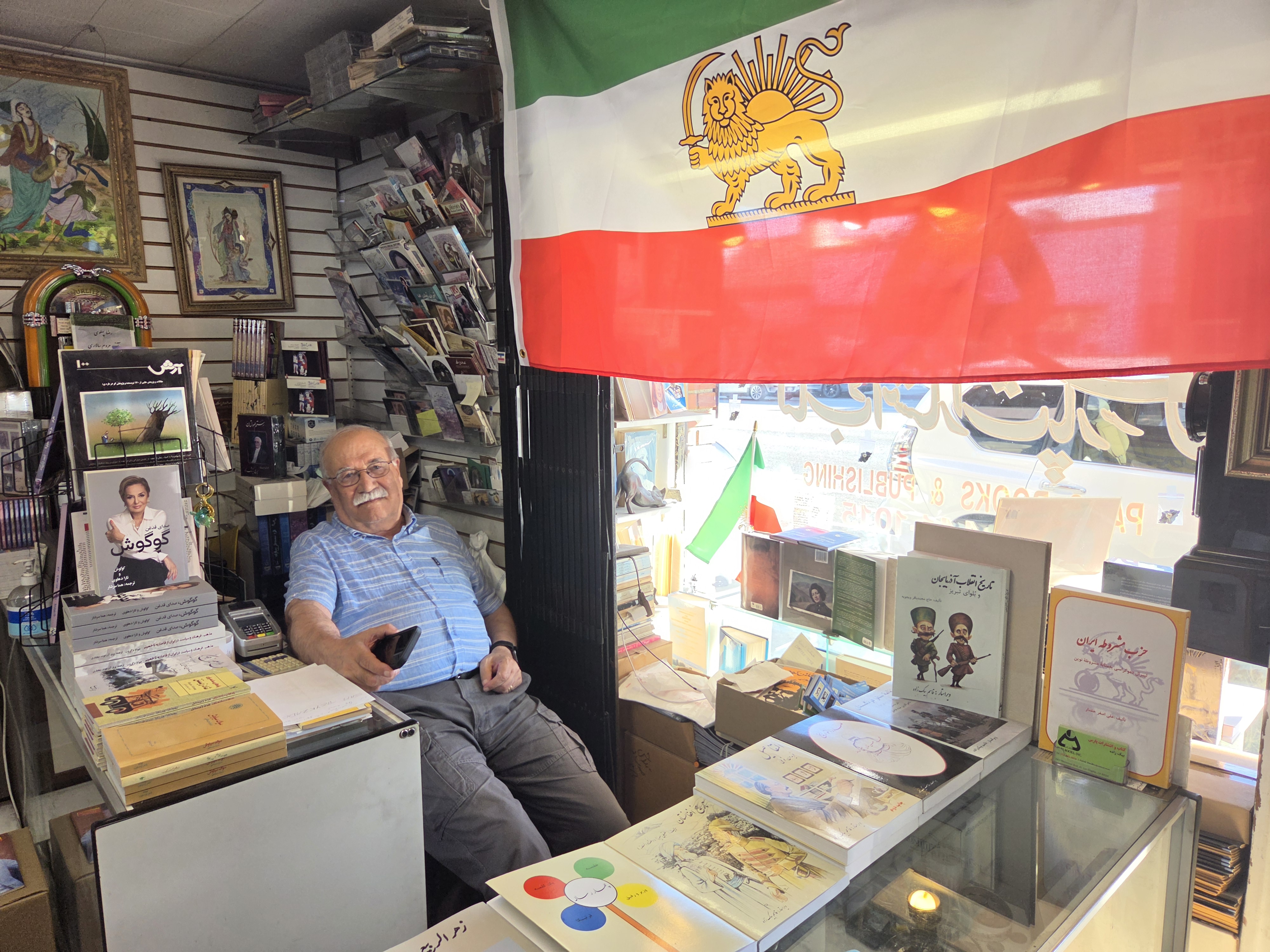 Man sits in a bookshop by a green, white and red flag with a drawing of a lion in the middle.