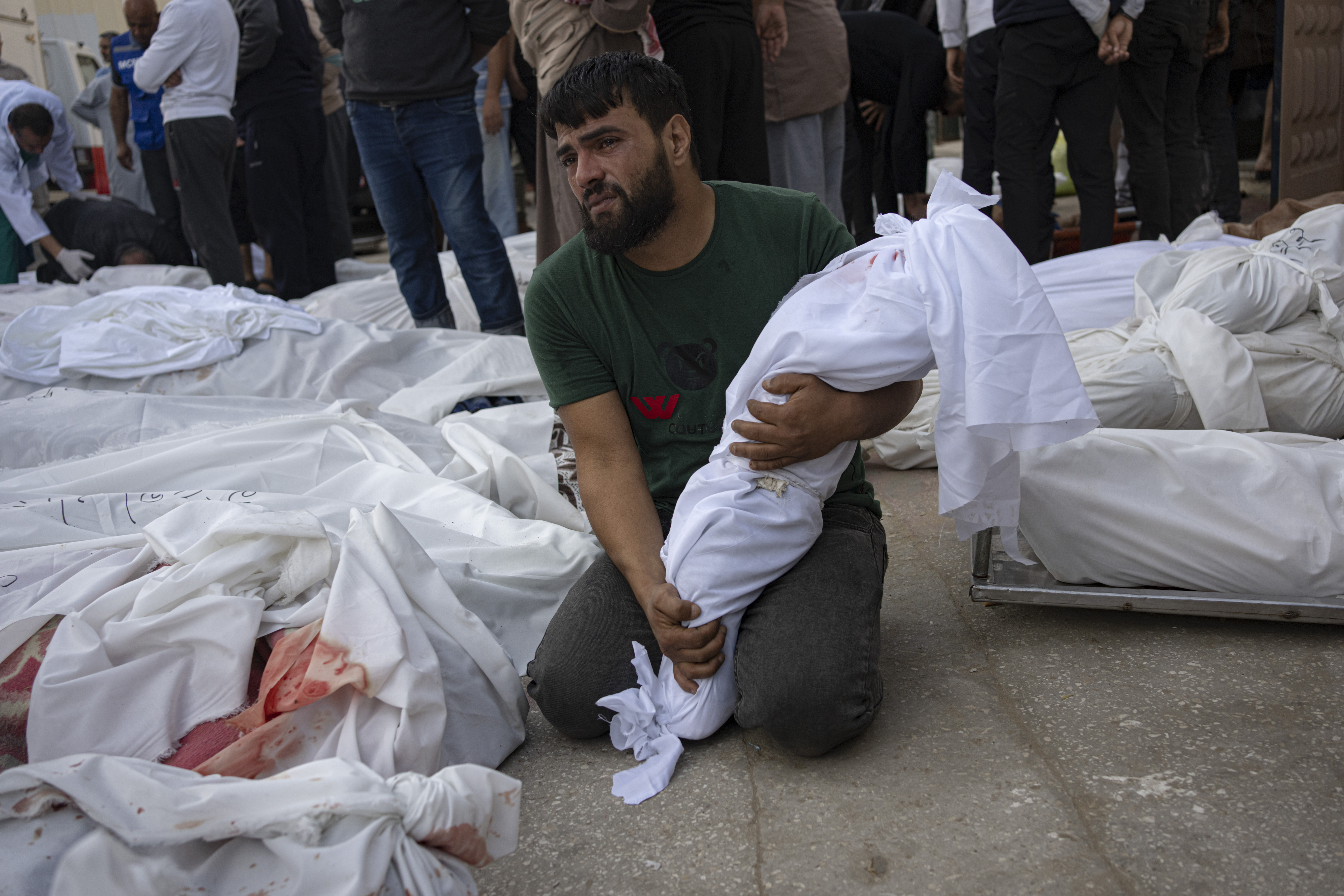 A man holds a small body bag next to other bodies in body bags on the ground.
