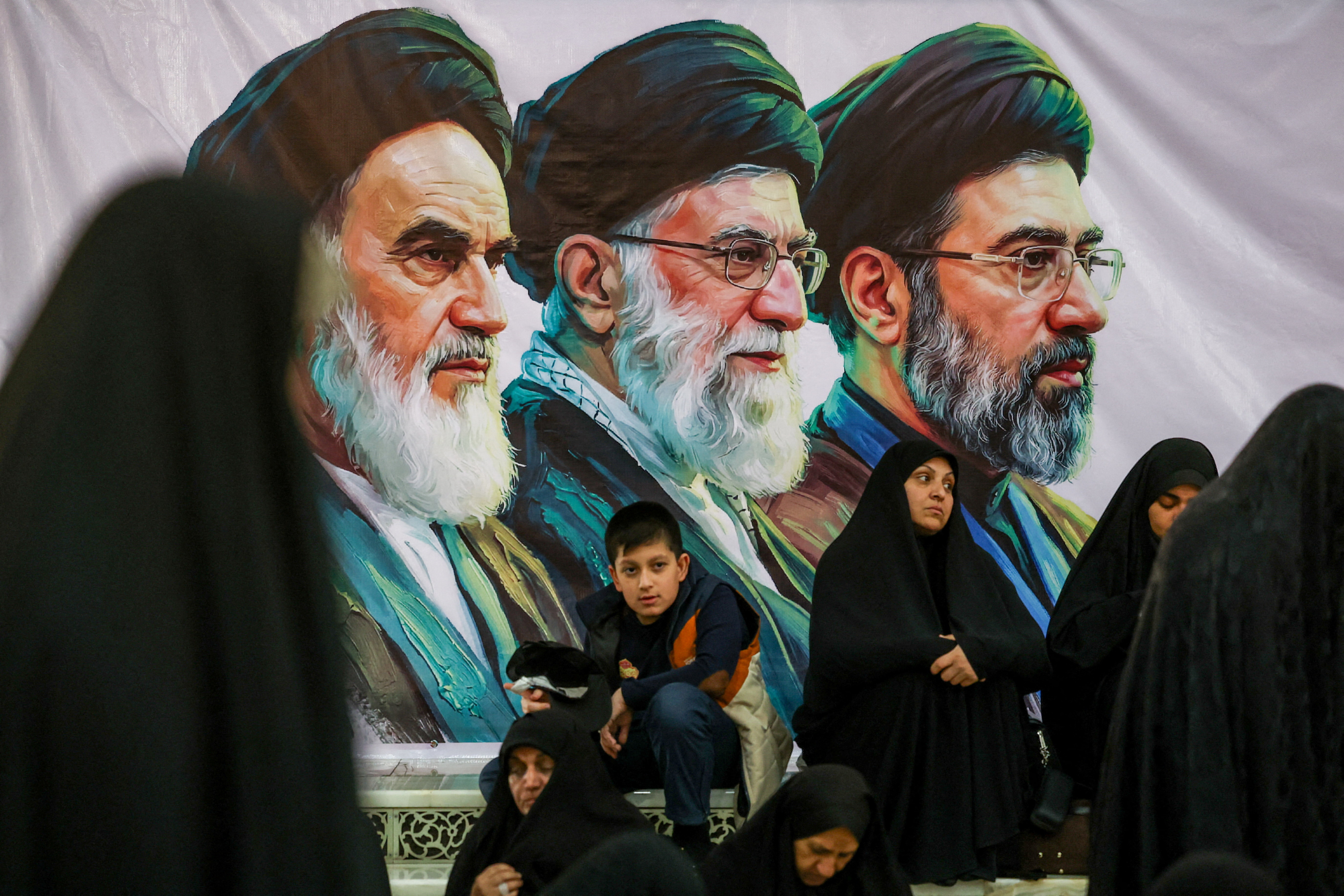 People gather near a banner with images of Iran's late leader of the Islamic revolution Ayatollah Ruhollah Khomeini, Iran's late Supreme Leader Ayatollah Ali Khamenei and Iran's Supreme Leader Mojtaba Khamenei.