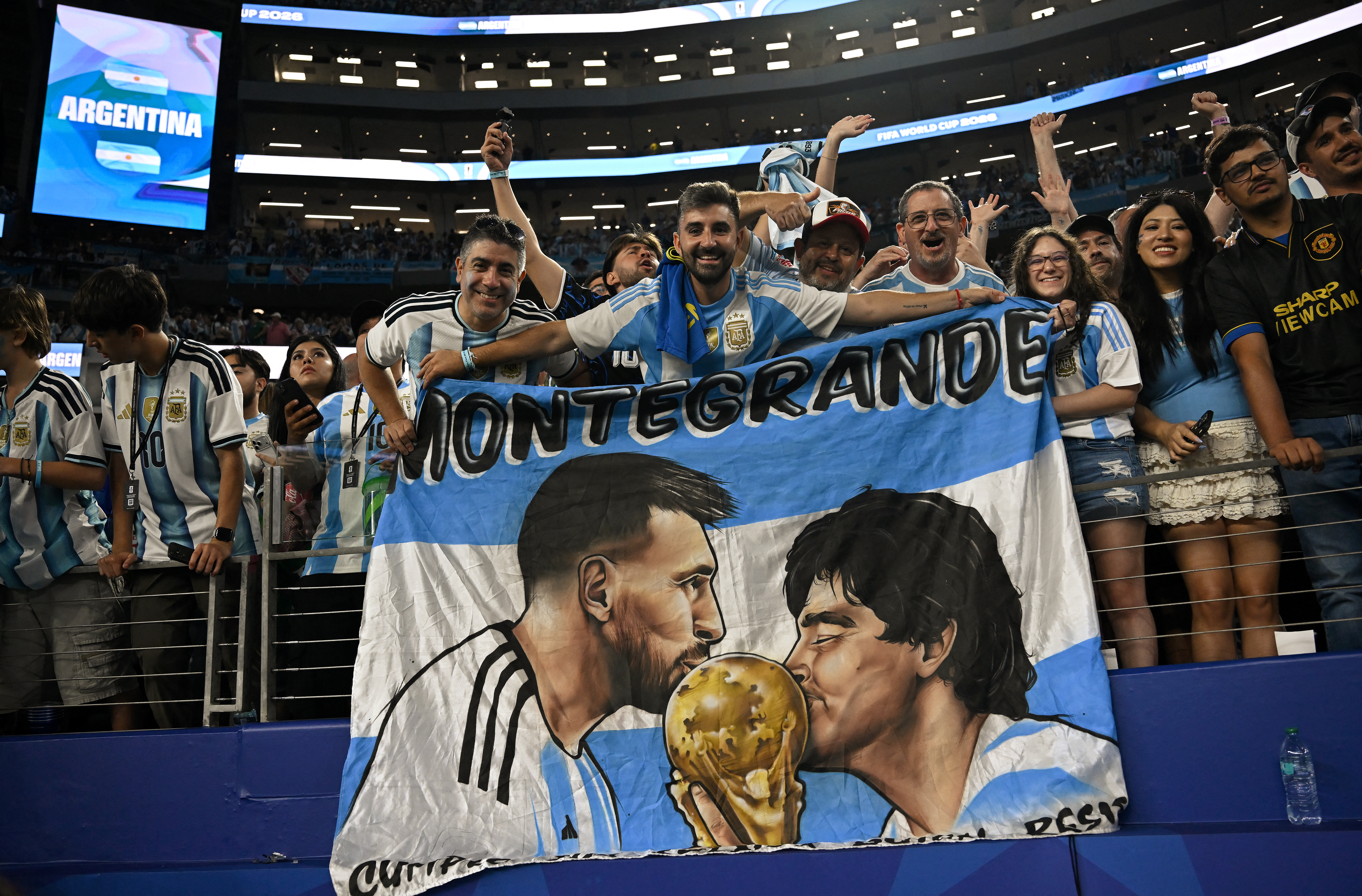 Argentine fans display a banner depicting Argentine football players Lionel Messi and Diego Maradona kissing the World Cup trophy as they celebrate at the end of the 2026 World Cup Group J football match between Jordan and Argentina at the Dallas Stadium in Arlington on June 27, 2026. (Photo by Paul ELLIS / AFP)