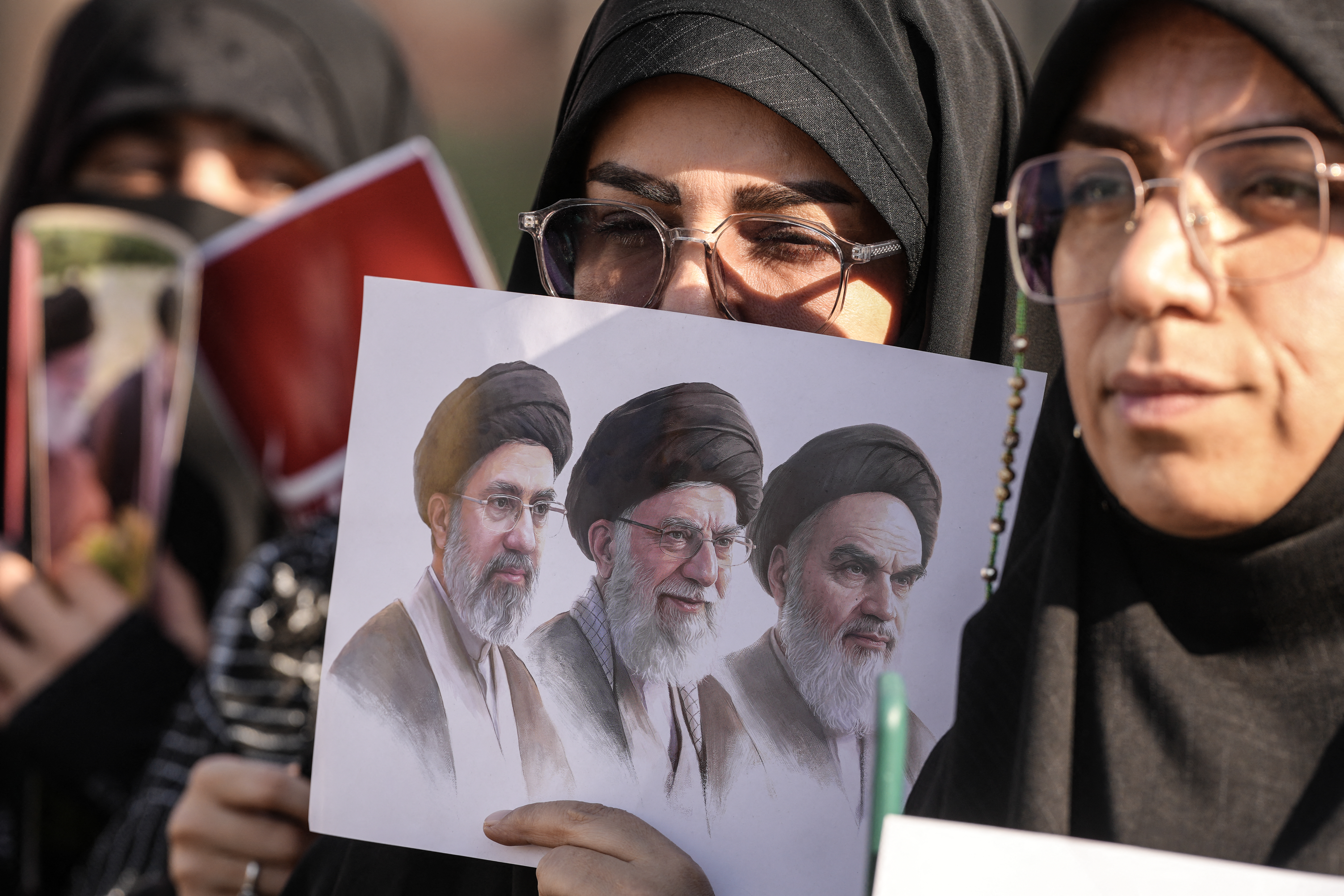 Mourners hold a placard depicting (from R) the late founder of the Islamic Republic, Ayatollah Ruhollah Khomeini, slain supreme leader Ayatollah Ali Khamenei, and current supreme leader Mojtaba Khamenei during a funeral procession for Ayatollah Ali Khamenei in Najaf on July 8, 2026. Crowds thronged the streets of Najaf on July 8 as the coffin of Khamenei moved through the holy city in a procession devoted to Iraq, home to Shia Islam's most sacred shrines. Iran began six days of public funeral ceremonies for Khamenei on July 4, including a dedicated day to neighbouring Iraq, a Shia powerhouse with close ties to Tehran. (Photo by Qassem al-KAABI / AFP)