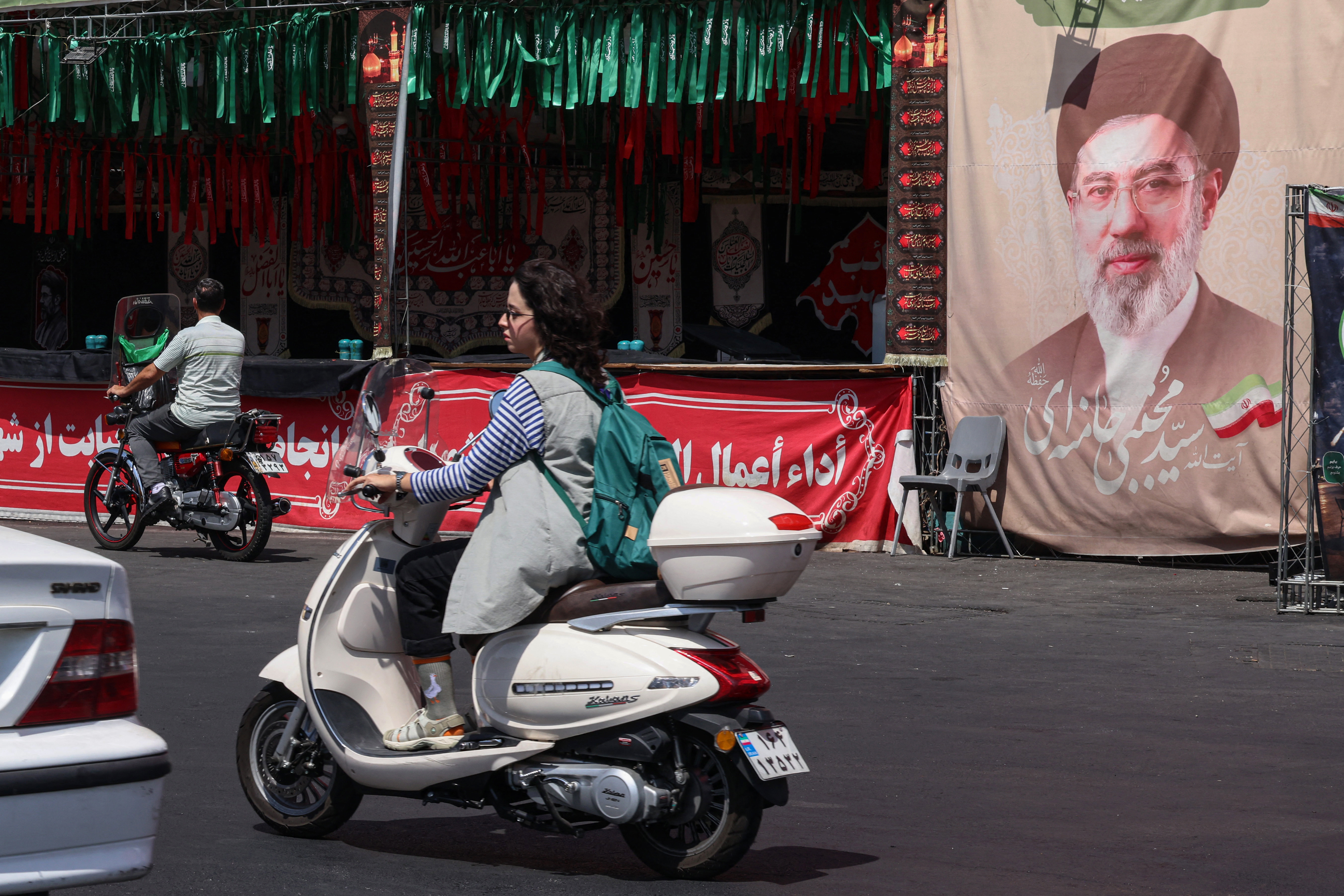 A woman rides a motorcycle past a banner of Iran's Supreme Leader Mojtaba Khamenei at Valiasr Square in Tehran on July 19, 2026. The United States said airstrikes on July 19 aimed to "punish" Iran over the first US military deaths since renewed hostilities with the Islamic republic. (Photo by ATTA KENARE / AFP) /
