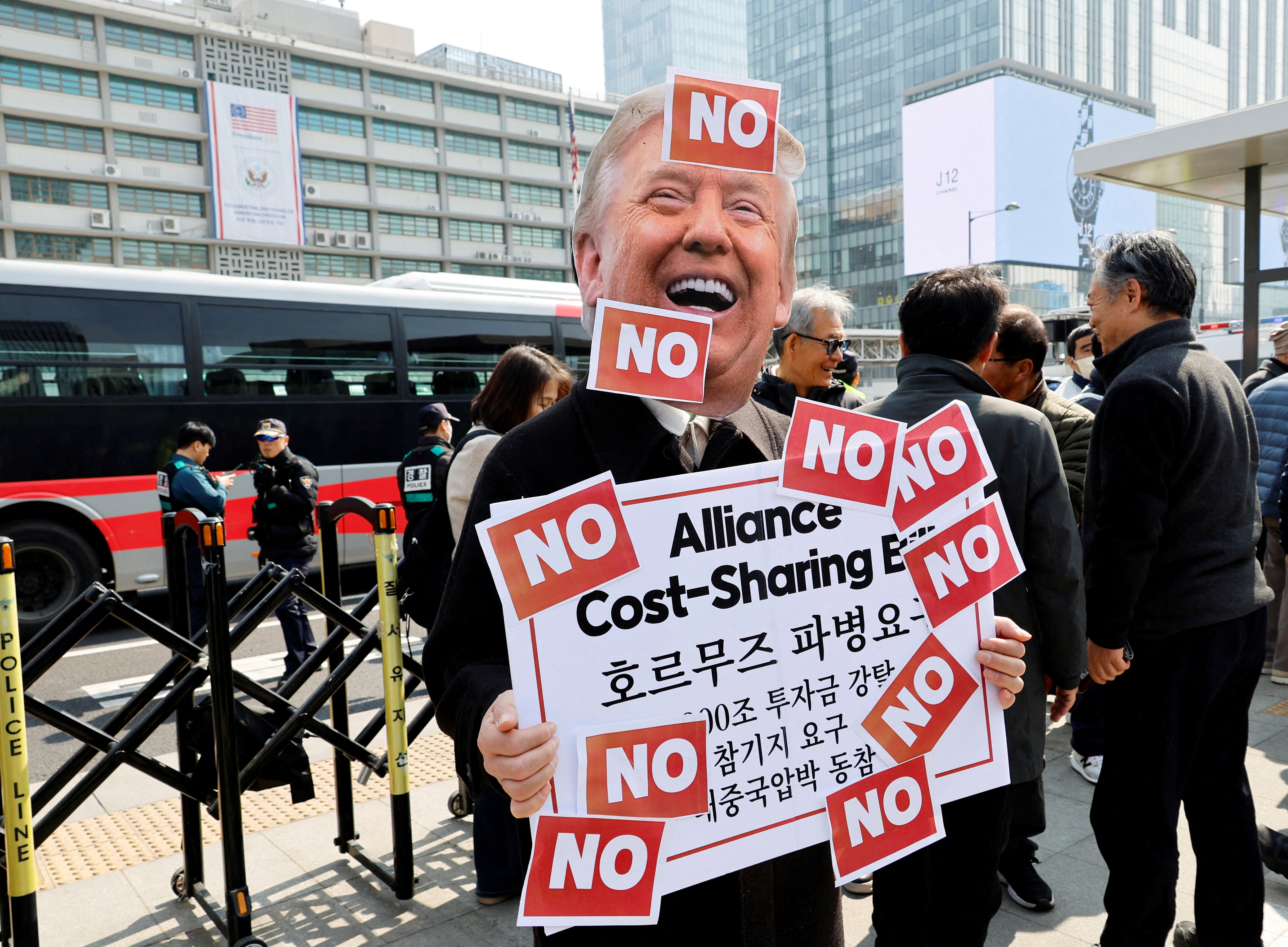 An activist wearing a cutout mask depicting U.S. President Donald Trump holds a placard covered with "No" stickers during a rally against the U.S. demand for South Korea to deploy troops to the Strait of Hormuz, outside the U.S. embassy in Seoul, South Korea, March 16, 2026.