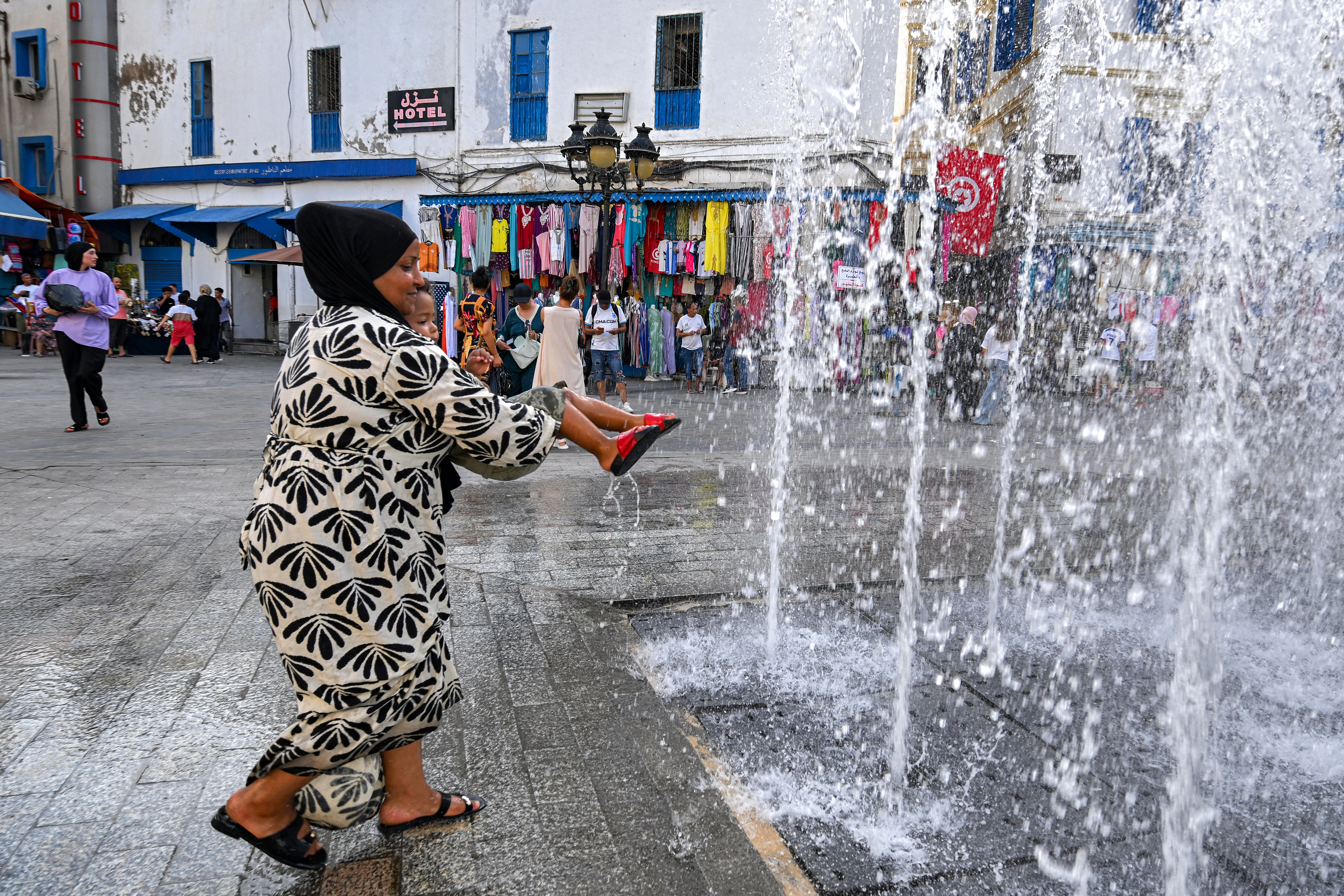 A woman carries a child as they cool off by water jets from a fountain at the entrance to the Medina in Tunis on August 24, 2026, amid a heatwave with temperatures exceeding 48°C and daily water and electricity cuts [Fethi Belaid/AFP]