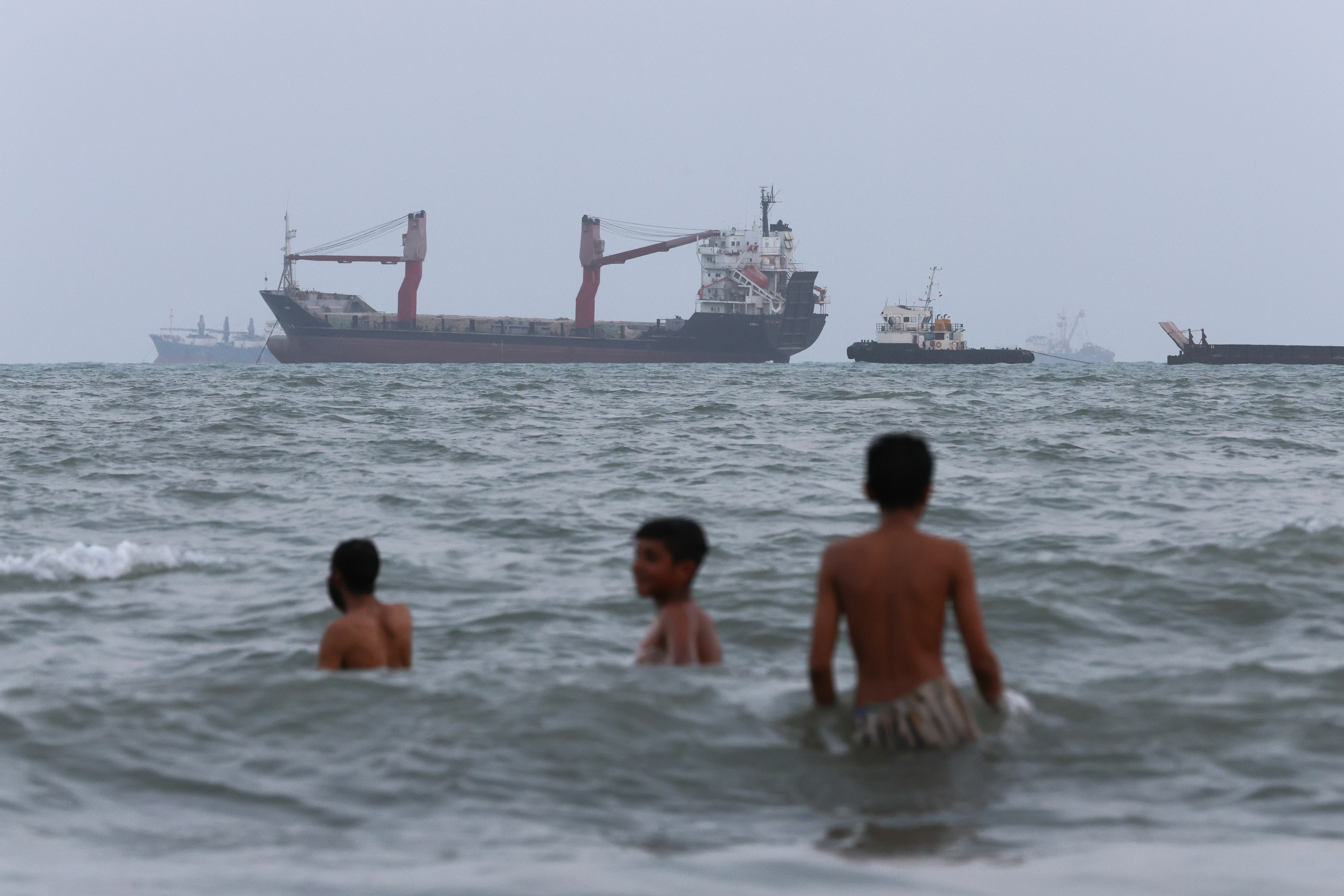 Children swim as ships are seen anchored in the Strait of Hormuz on August 10, 2026, off the coast of Bandar Abbas, Iran