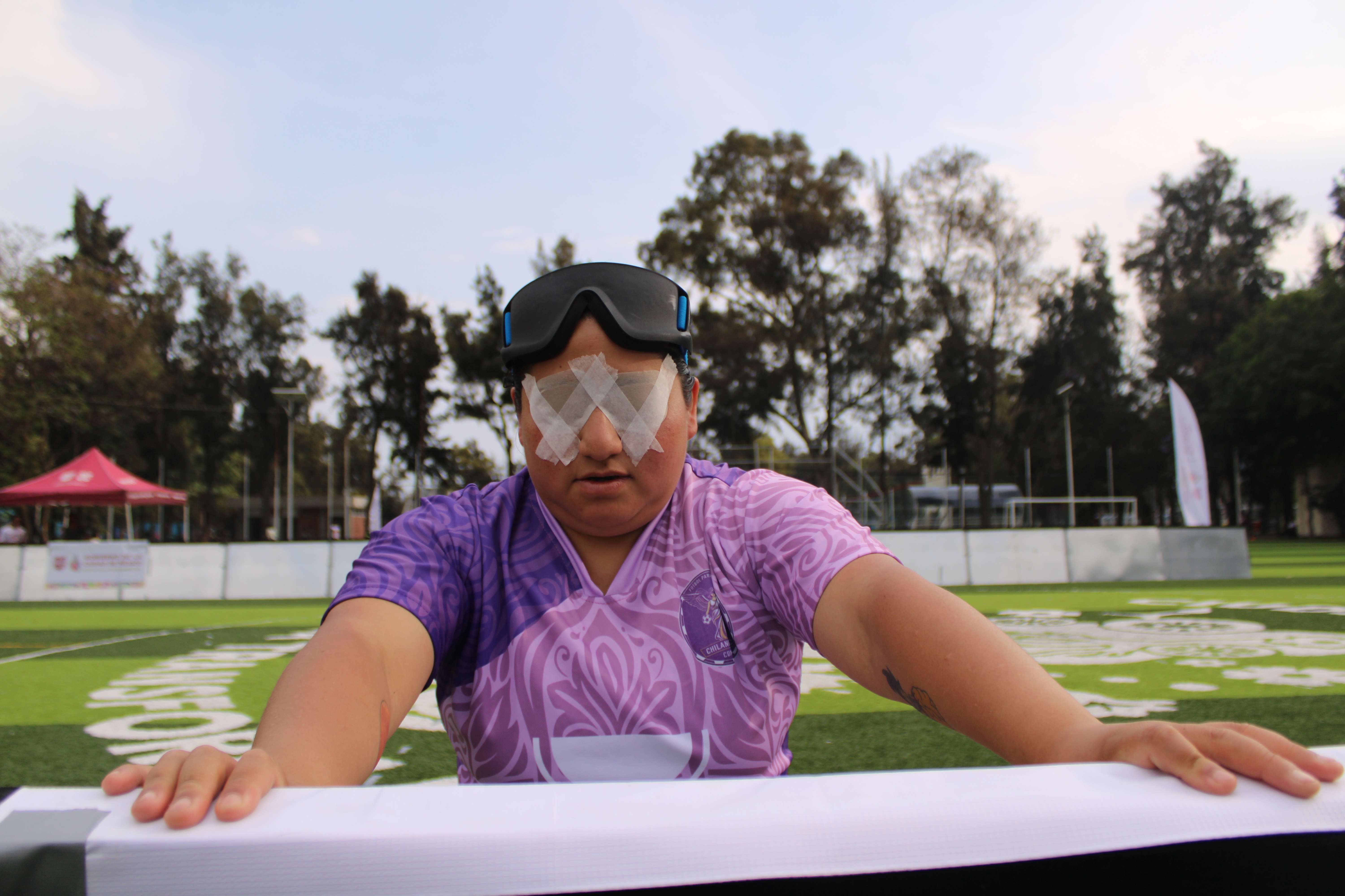 Pau prepares for a match during a national blind soccer tournament in Mexico City, leaning against the boards that surround the pitch as she lifts her visor to reveal protective eye patches secured with athletic tape. In official blind soc