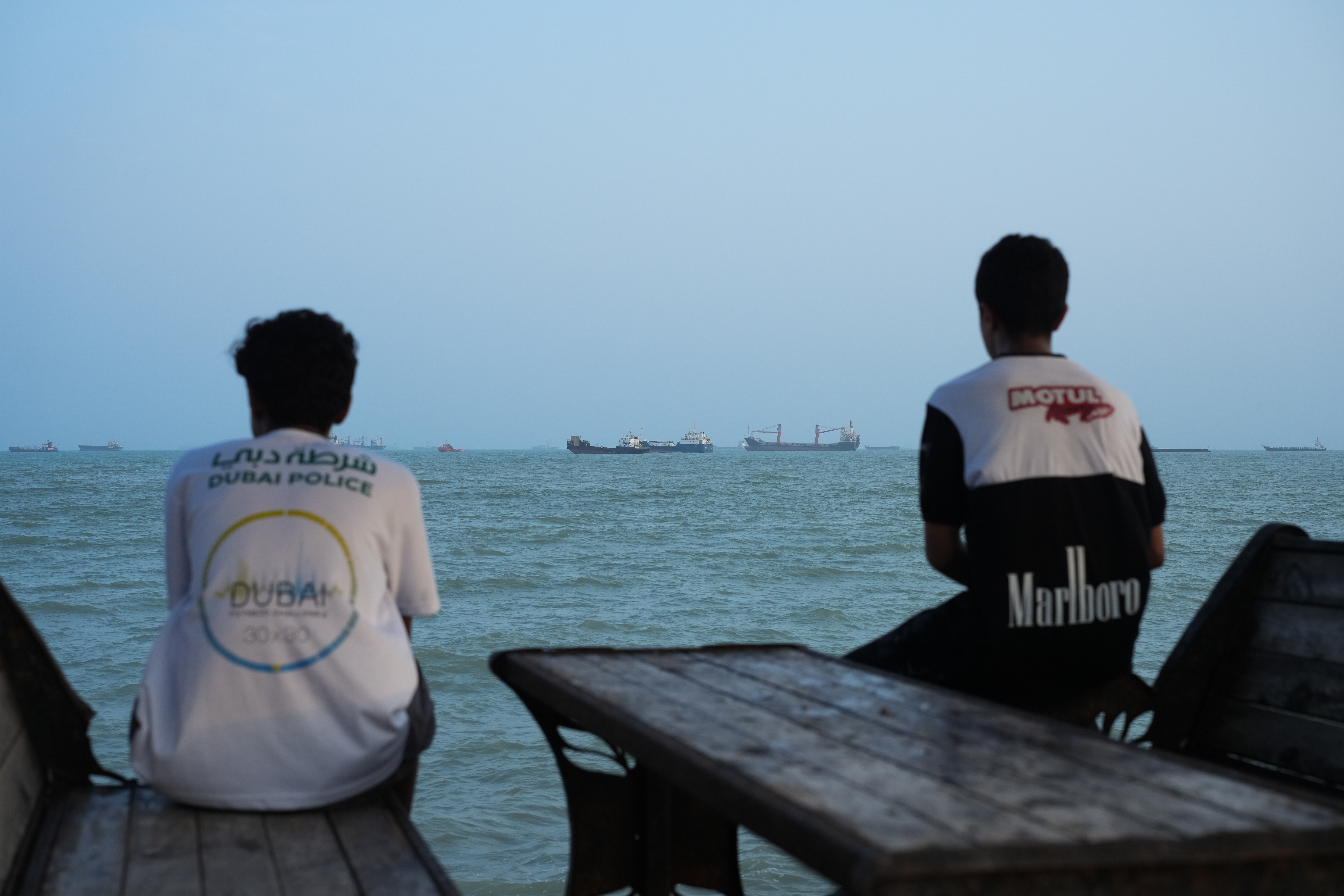 Two youths sit on the shoreline as ships are seen anchored in the Strait of Hormuz on August 10, 2026, off the coast of Bandar Abbas, Iran