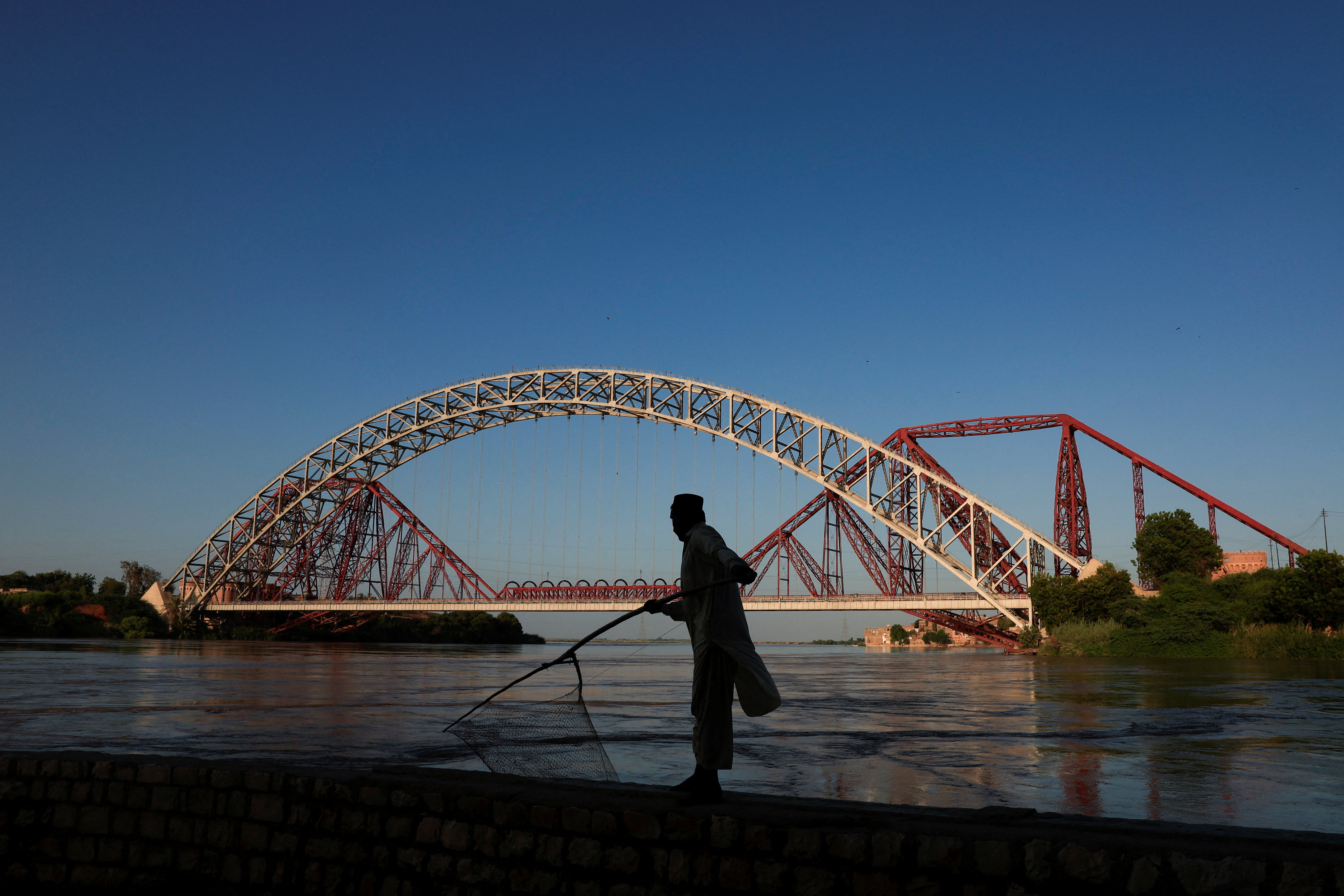Ali Muhammad, 45, is silhouetted as he walks with a fish net on a wall, with the Lansdowne Bridge that was built between 1887 and 1889, during the British Raj era, in the background, as floodwater passes through, following monsoon rains and rising levels of the Indus River in Sukkur, Sindh province, Pakistan September 13, 2025. REUTERS/Akhtar Soomro